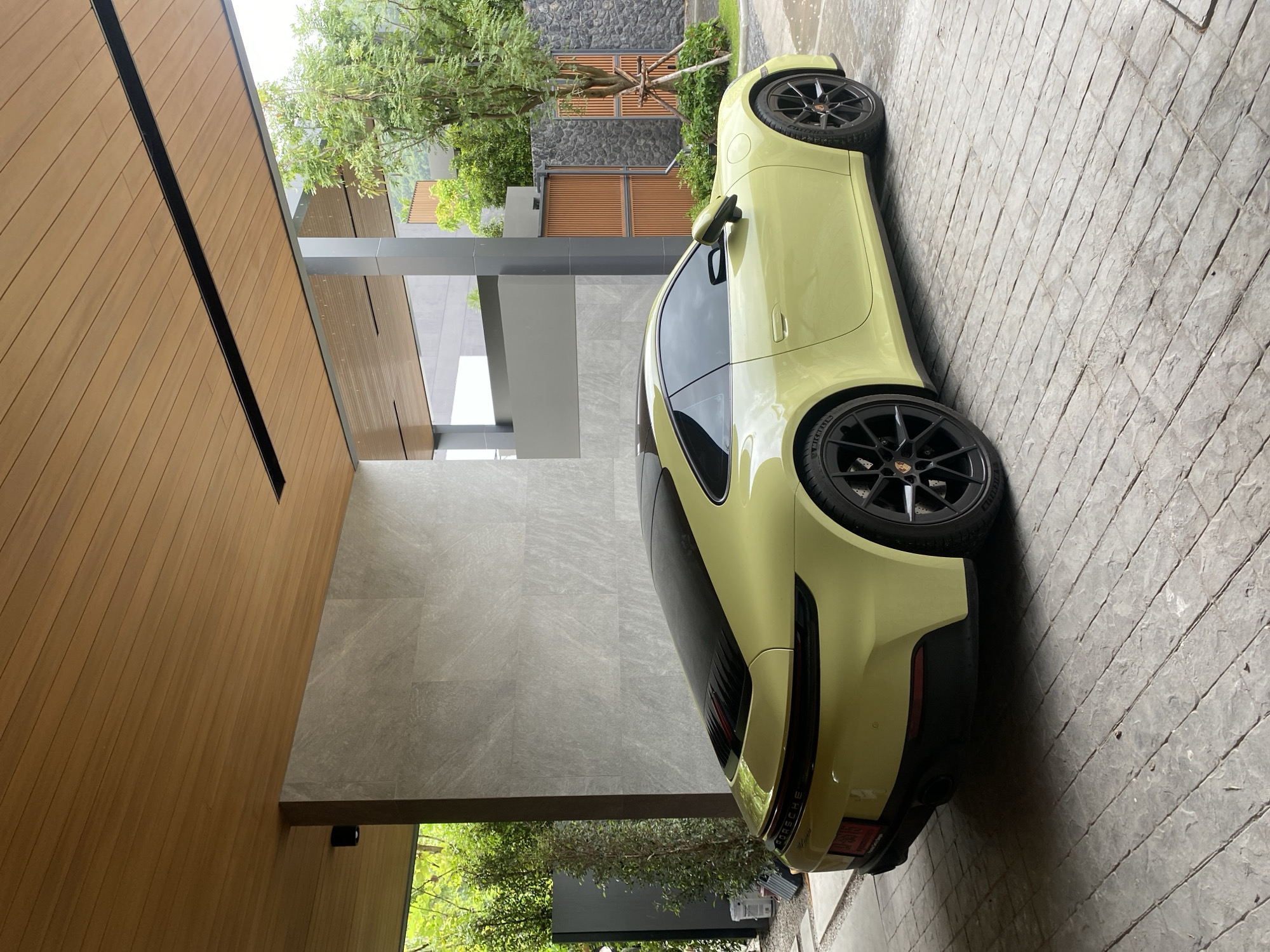 A light yellow Porsche coupe is parked on a gray stone driveway next to a modern building, likely a private residence, in Choeng Thale, Thailand. The building features a combination of wood paneling, gray stone, and an open entryway with greenery visible. The car has black wheels and its rear lights are visible. The scene suggests a well-maintained, upscale residential area. There are no visible people.