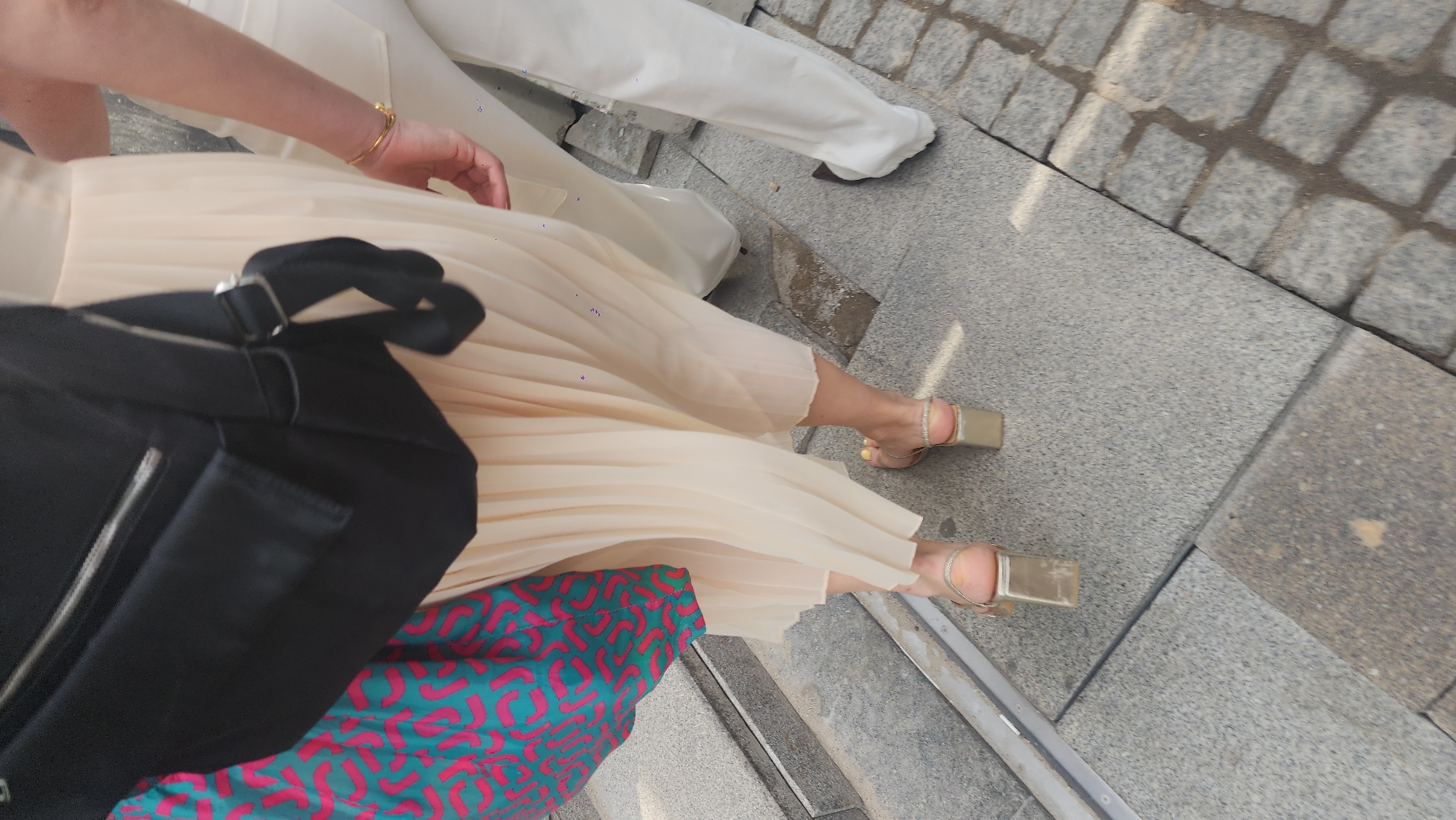 The image captures a scene from a high-angle perspective, likely taken in an outdoor urban environment. The lower legs and feet of two individuals are visible. One person is wearing white trousers and the other is wearing a pleated, light-colored dress, adorned with subtle shimmering details. The person in the dress also has gold-colored high-heeled sandals with ankle straps. A large black shoulder bag obscures part of the dress and a colorful, patterned fabric is partially visible beneath. The setting appears to be a pedestrian area, judging by the gray stone paving. The image does not offer sufficient detail to determine the specific location in Moscow, Russia.