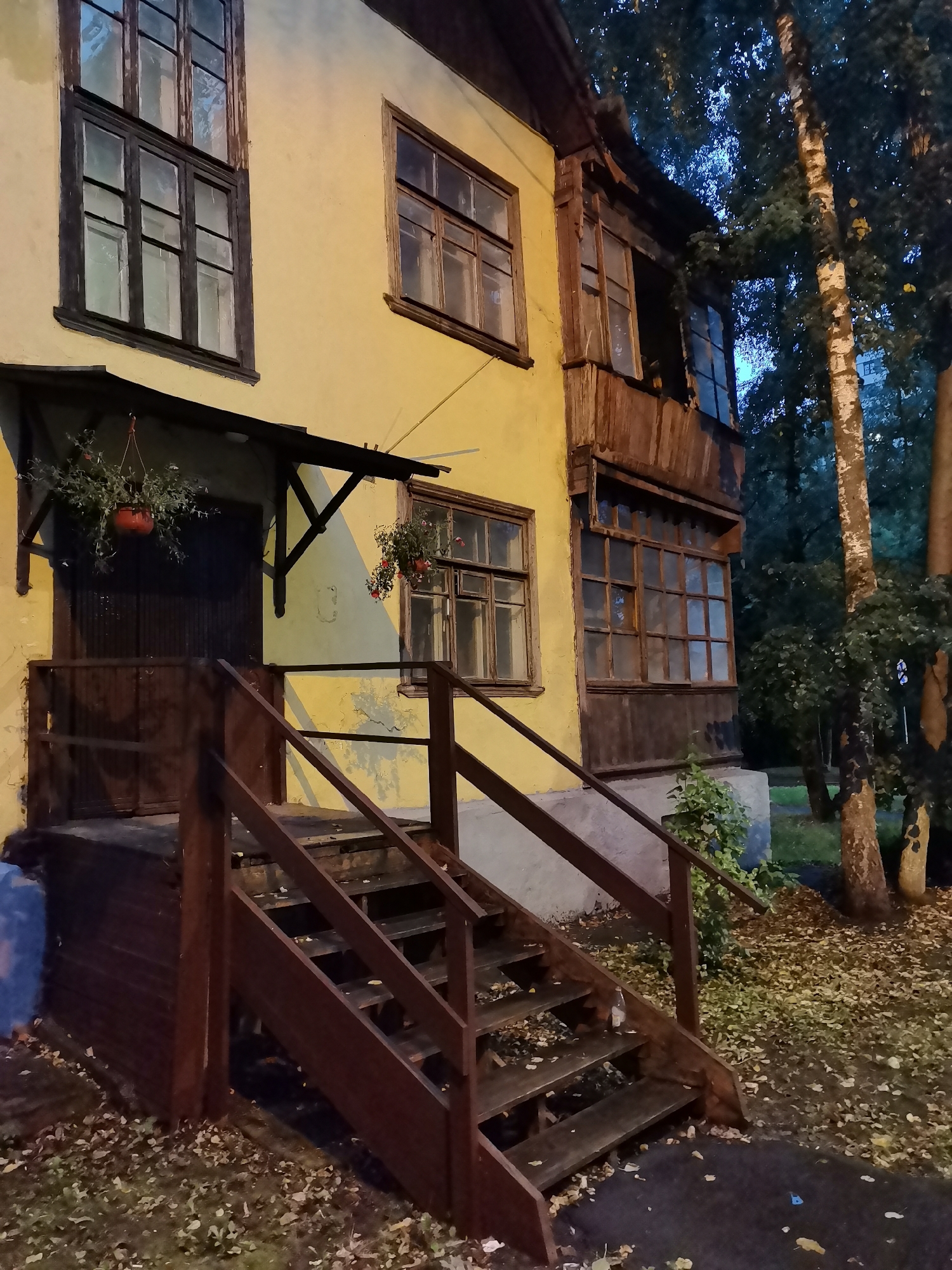A dimly lit, two-story yellow building with dark wooden window frames and a covered porch is shown at dusk. A wooden staircase with a handrail leads up to the porch. Potted plants hang from the porch roof and near a window. The building is surrounded by trees and fallen leaves on the ground. The scene appears to be in a residential area.