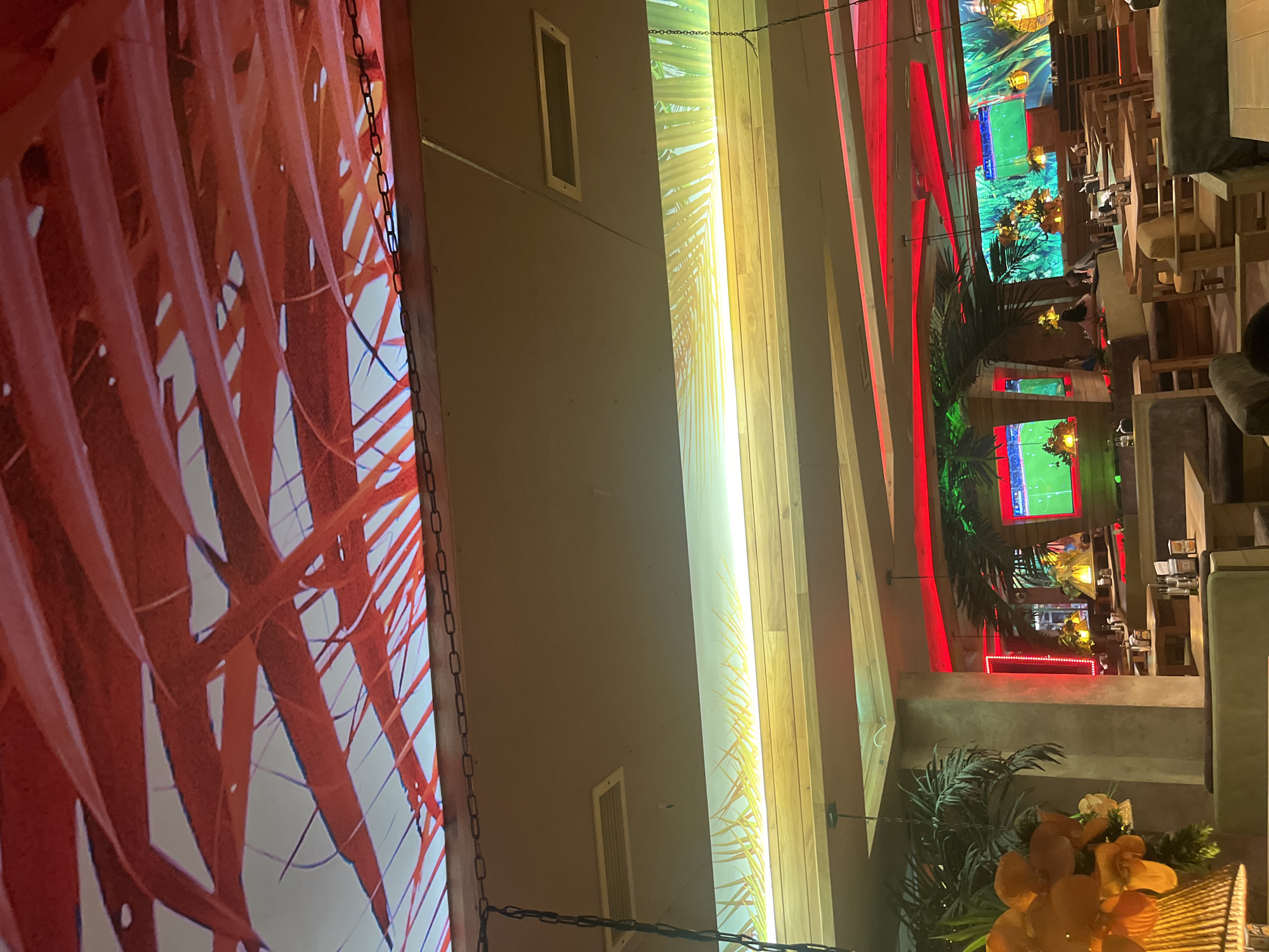 The interior of a restaurant in Sofia, Bulgaria. A large screen with an image of red leaves is on the left. There is a tan wall with vents in the center. Yellow and white lights in wooden slats illuminate the space. The right side features seating areas with green palm trees and a decorative orange floral arrangement. Screens on the back wall show scenes of foliage and soccer matches. Red lights accent the ceiling and architectural features. People are seated at tables.