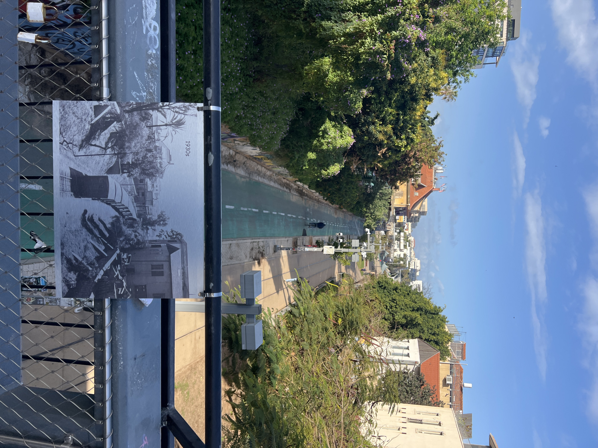 The image depicts a bridge overlooking a bike path in Tel Aviv, Israel. The bike path is lined with vegetation on either side and stretches into the distance. Along the sides of the path are buildings. A person on a bicycle is visible on the path. Affixed to the bridge railing is a black and white photograph dated "1930s". The sky is blue with scattered clouds.