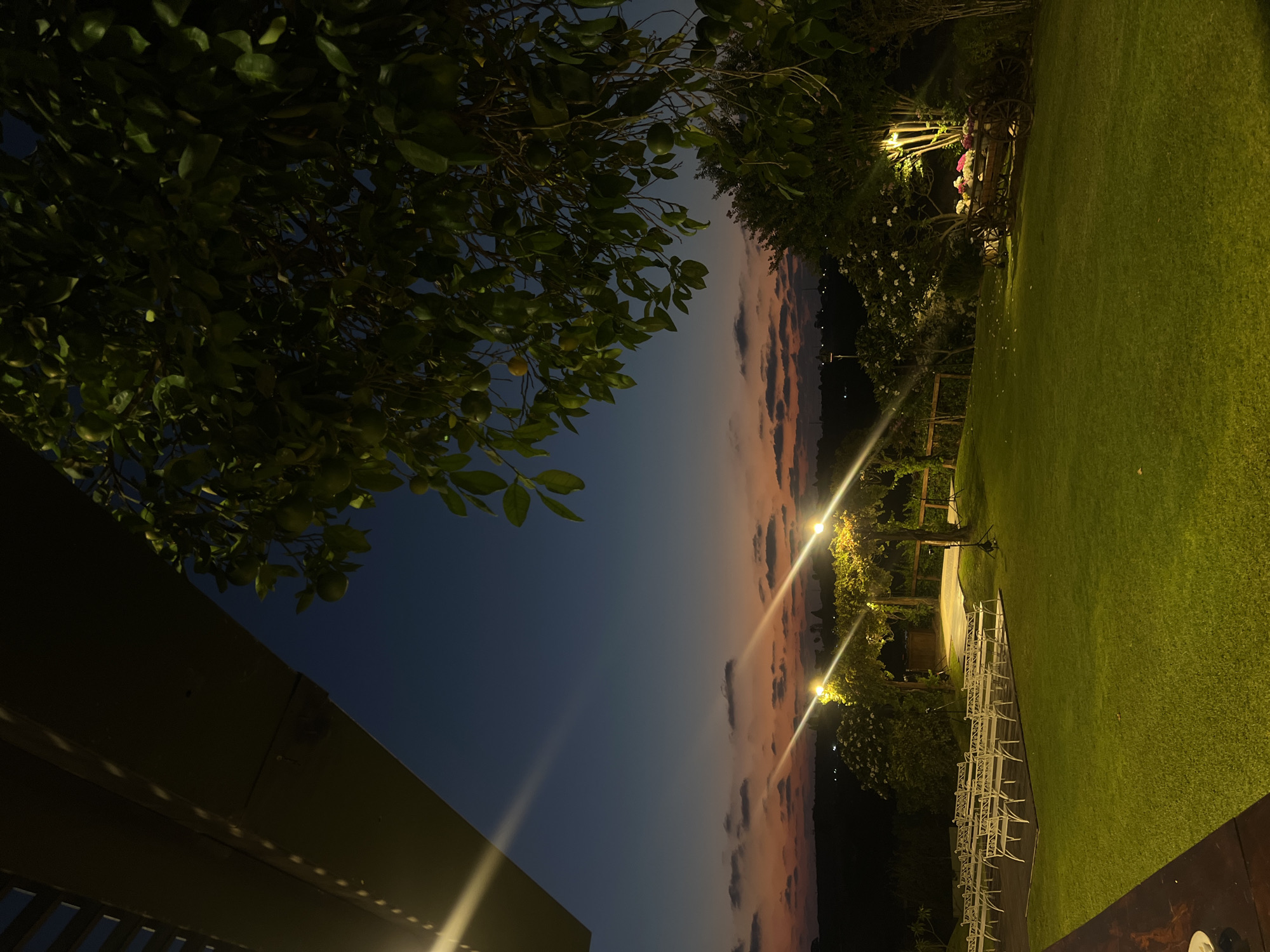 The image shows an outdoor event space at dusk in Beit Hanan, Israel. Rows of white chairs are set up on a grassy lawn, facing a wooden platform adorned with string lights. A tree with green leaves and fruit is in the foreground on the right, and another tree with white flowers is visible behind the chairs. The sky is a gradient of deep blue to orange, with scattered clouds. Lights are illuminating the scene. There are no people visible.