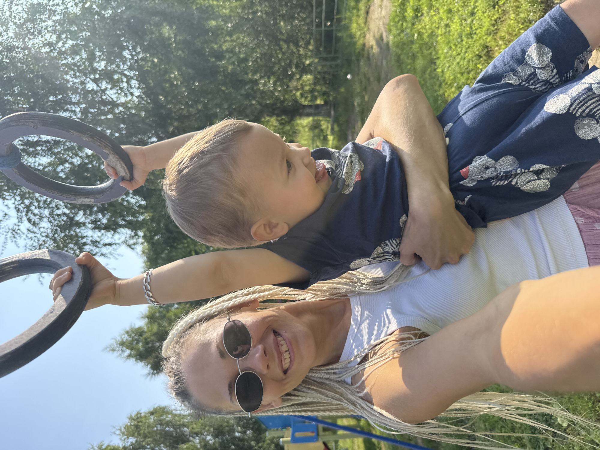 In Ignatovo, Russia, a woman with blonde braided hair and sunglasses smiles at the camera, holding a young boy in her arms. The boy, wearing a blue t-shirt and shorts with a teddy bear pattern, reaches up towards a ring suspended from above. They are outdoors on a sunny day, with green grass and trees in the background. A playground structure is visible to the left.