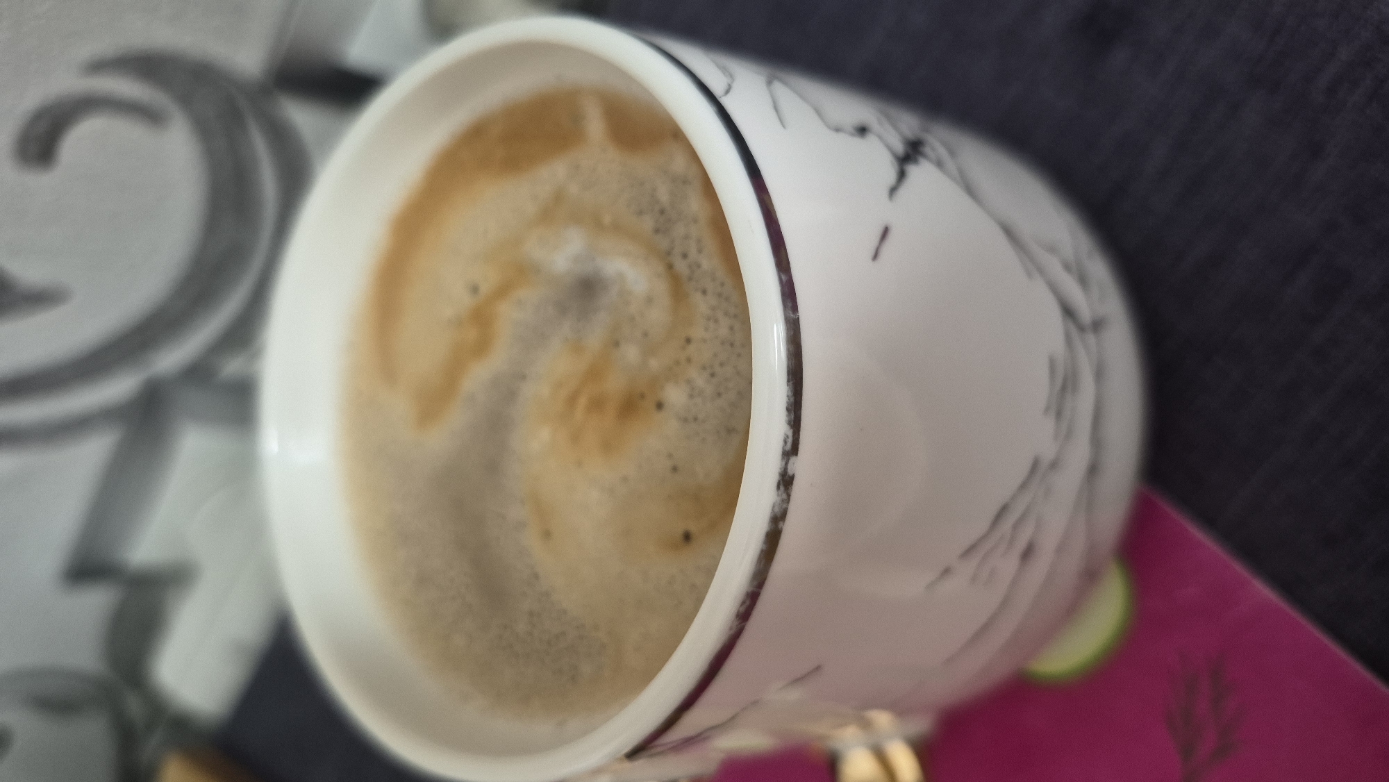 A white ceramic mug with a marble-like pattern and a thin silver rim contains coffee with a frothy head. The mug sits on a dark blue surface, possibly a table or placemat. To the left of the mug, a portion of a bright pink object with a green circular detail is visible, possibly a coaster or part of a placemat. The background is out of focus, showing what appears to be a textured wall with a gray, swirling pattern. There is no indication of the city of Krauja, Latvia, in the image.