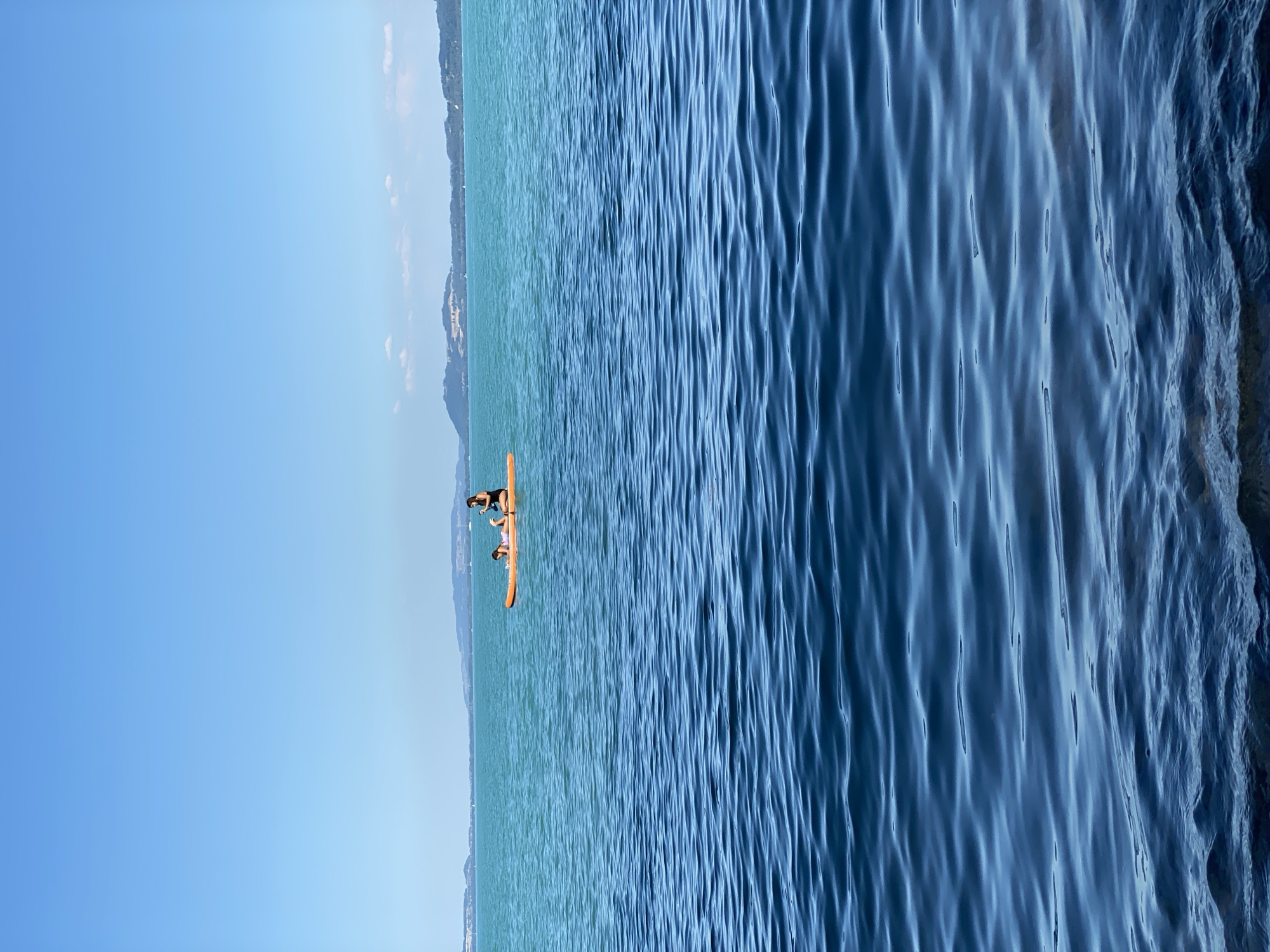 Two individuals are on an orange paddleboard in the turquoise waters of Lake Neuchâtel, near Cortaillod, Switzerland. The person in the back is kneeling and appears to be guiding the board, while the person in the front is lying down. The water is calm, with gentle ripples. In the distance, the hazy outlines of hills and mountains can be seen on the horizon under a clear blue sky.