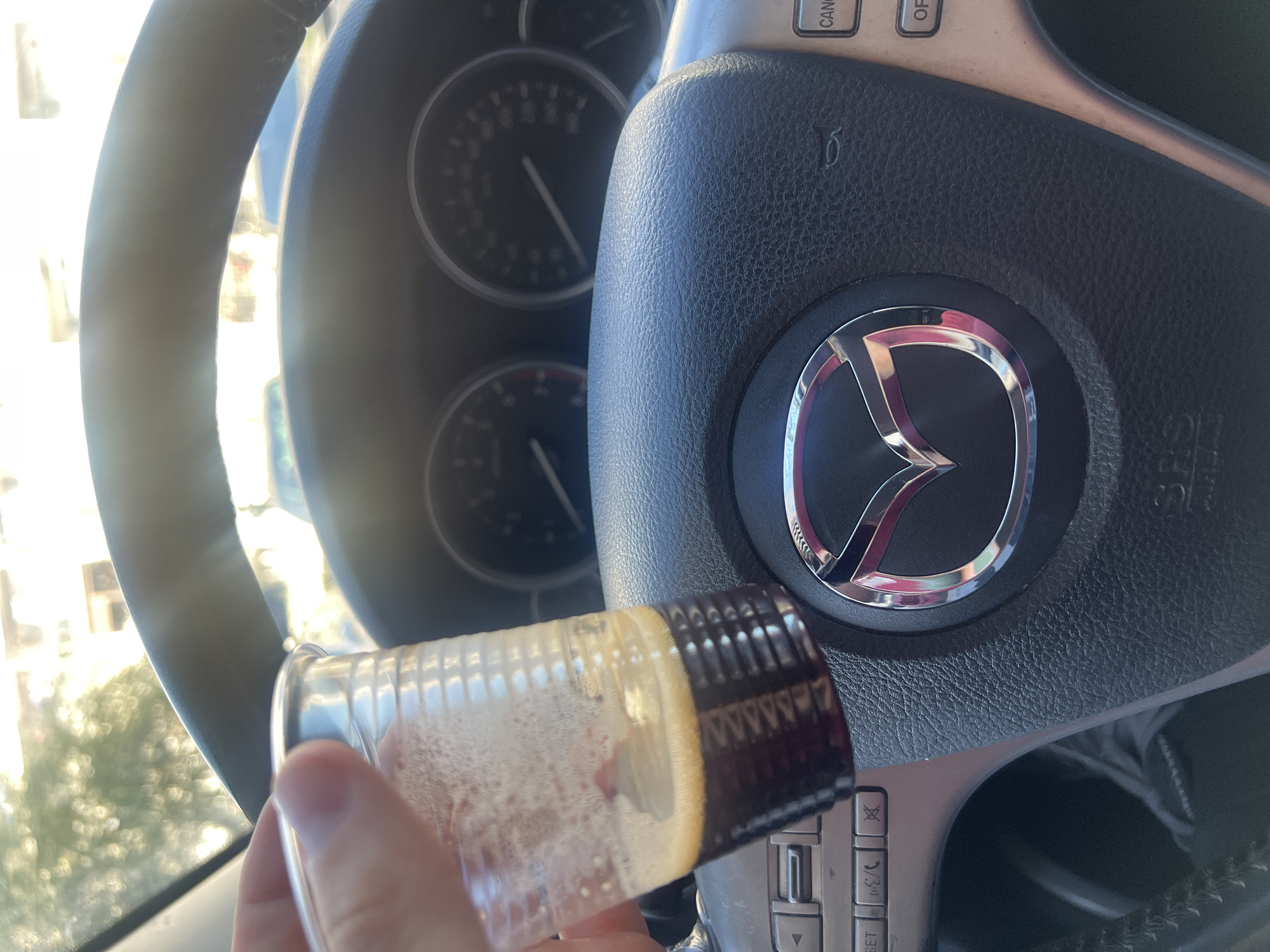 The image shows the interior of a Mazda car, specifically the steering wheel with the Mazda logo prominently displayed. A partially filled plastic cup, containing a reddish-brown liquid, is being held in front of the steering wheel. The instrument cluster is visible in the background, though details are somewhat obscured by glare and depth of field. The environment appears to be outdoors, indicated by light coming through the windshield and trees/buildings visible in the background. Given the request to include location in Burgas, Bulgaria (although it is not possible to confirm this from the photograph), it is inferred that this image was taken in Burgas.