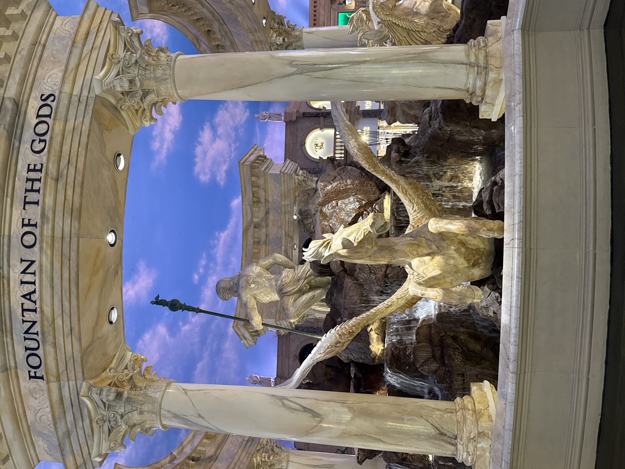 The image shows the "Fountain of the Gods" located in the Forum Shops at Caesars Palace on the Las Vegas Strip in Paradise, Nevada, United States of America. The fountain features several sculptures in a classical Greco-Roman style. In the foreground, a winged horse, likely Pegasus, is depicted in mid-leap above a rocky cascade. Behind Pegasus, a muscular statue of a male figure, possibly a god like Neptune or Poseidon, kneels while holding a trident. Water flows down the rocks, creating a waterfall effect. The fountain is framed by large, ornate columns and an arched ceiling painted with a sky motif. The overall scene evokes a sense of grandeur and mythology, consistent with the opulent themed environments found in Las Vegas.