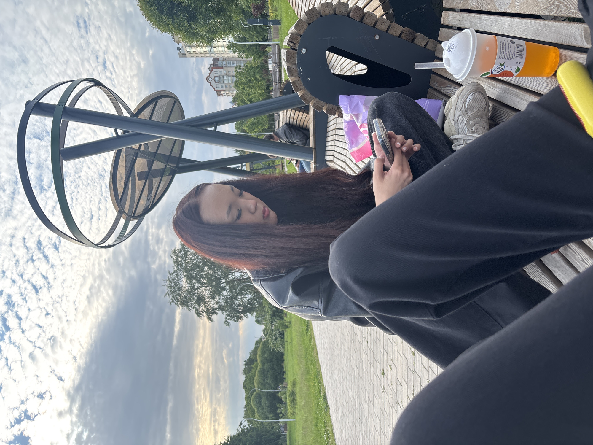 A young woman with long, dark brown hair sits on a park bench in Sinyavino, Russia, looking down at her mobile phone. She is wearing a black leather jacket and dark pants. To her right, on the same bench, is a to-go cup filled with an orange beverage and a straw. In the background, a modern-looking metal sculpture with circular elements stands against a cloudy sky. Behind the sculpture, trees and apartment buildings are visible. Another person is seated on a bench further in the background, also looking at their phone. The bench she is sitting on is made of wood with metal supports and has a decorative, carved edge. A patterned shopping bag is partially visible next to her on the bench. The ground in front of the bench is paved with interlocking bricks.