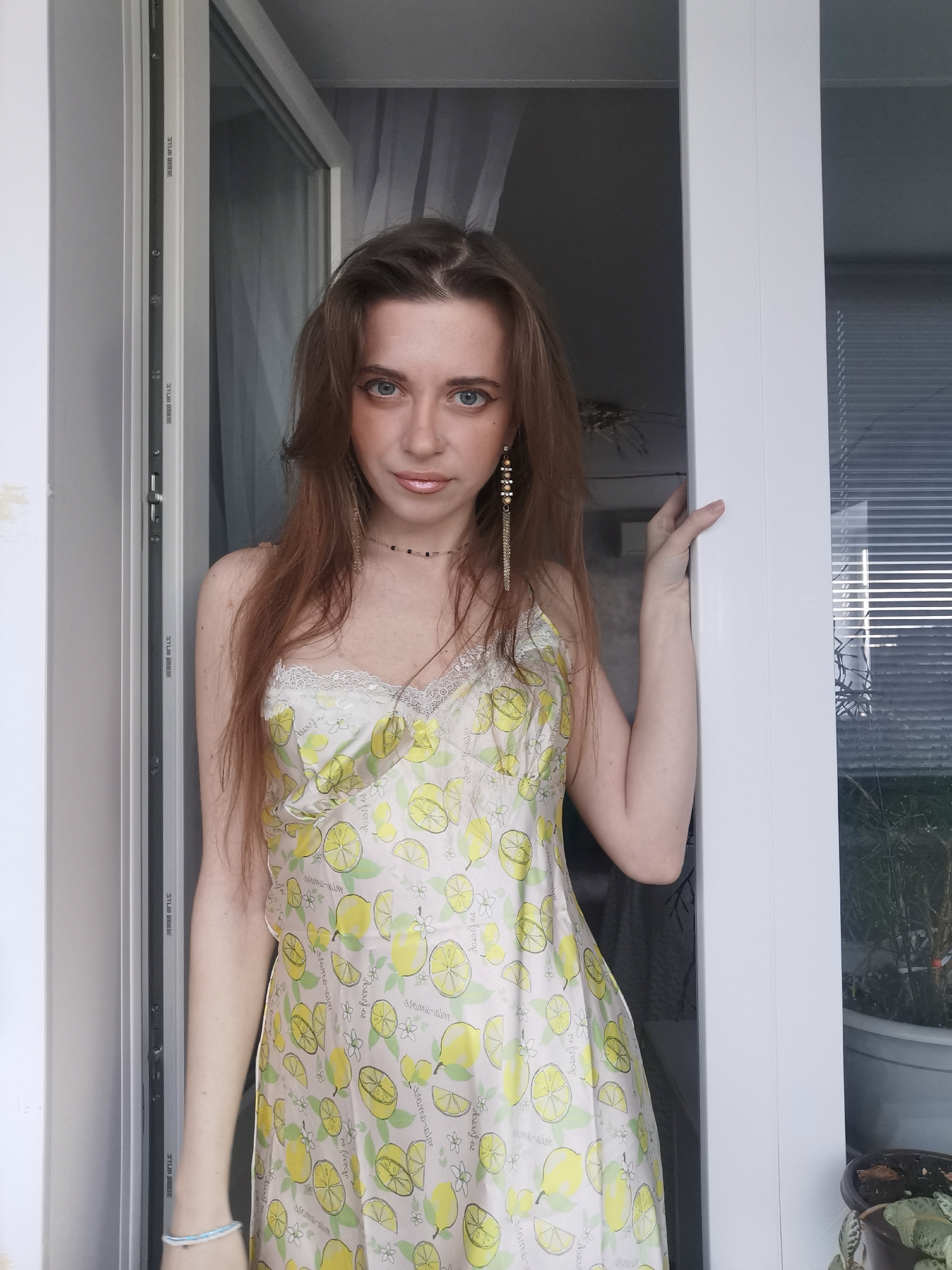 A young woman with long, reddish-brown hair and blue eyes is captured in a vertical, waist-up shot, looking directly at the viewer. She is standing in what appears to be a doorway or opening, holding onto the white frame with her right hand. She wears a light-colored, possibly white or cream, slip dress adorned with a vibrant pattern of yellow lemons, lemon slices, and small white flowers, complemented by white lace trim on the bodice. Long, dangling earrings with gold and dark beads are visible, along with a thin choker necklace and a blue and white beaded bracelet on her left wrist.
The background shows a partially open door or window, revealing internal blinds and some potted greenery. The overall setting appears to be a brightly lit indoor space, possibly a home. The woman has a neutral to slightly serious expression.
Visible text on the door frame, possibly reversed or embossed, includes "MEMAD", "ZULER OPTLS", and "STLLO DELLE". The fabric pattern on her dress also incorporates stylized, repeated text elements such as "wamo-alm", "assamo-nim", "Maru", "Brus", "ramoral", and "wbsang".