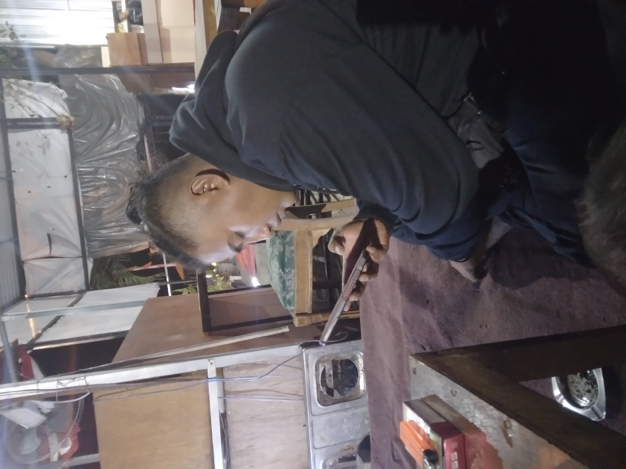 A person is seated on the ground, wearing a black hooded sweatshirt and dark pants. They are holding a mobile phone in their right hand and appear to be looking at it. The environment is outdoors, possibly a covered patio or an open-air structure. In the background, there are some structures with white sheeting, wooden panels, and furniture, including a chair with a green cushion. In the foreground, a wooden table is visible, with a cigarette pack and an orange lighter on it. An ashtray filled with cigarette butts is located underneath the table. The scene is dimly lit, suggesting it might be evening or nighttime. The location is identified as Pacet, Indonesia.