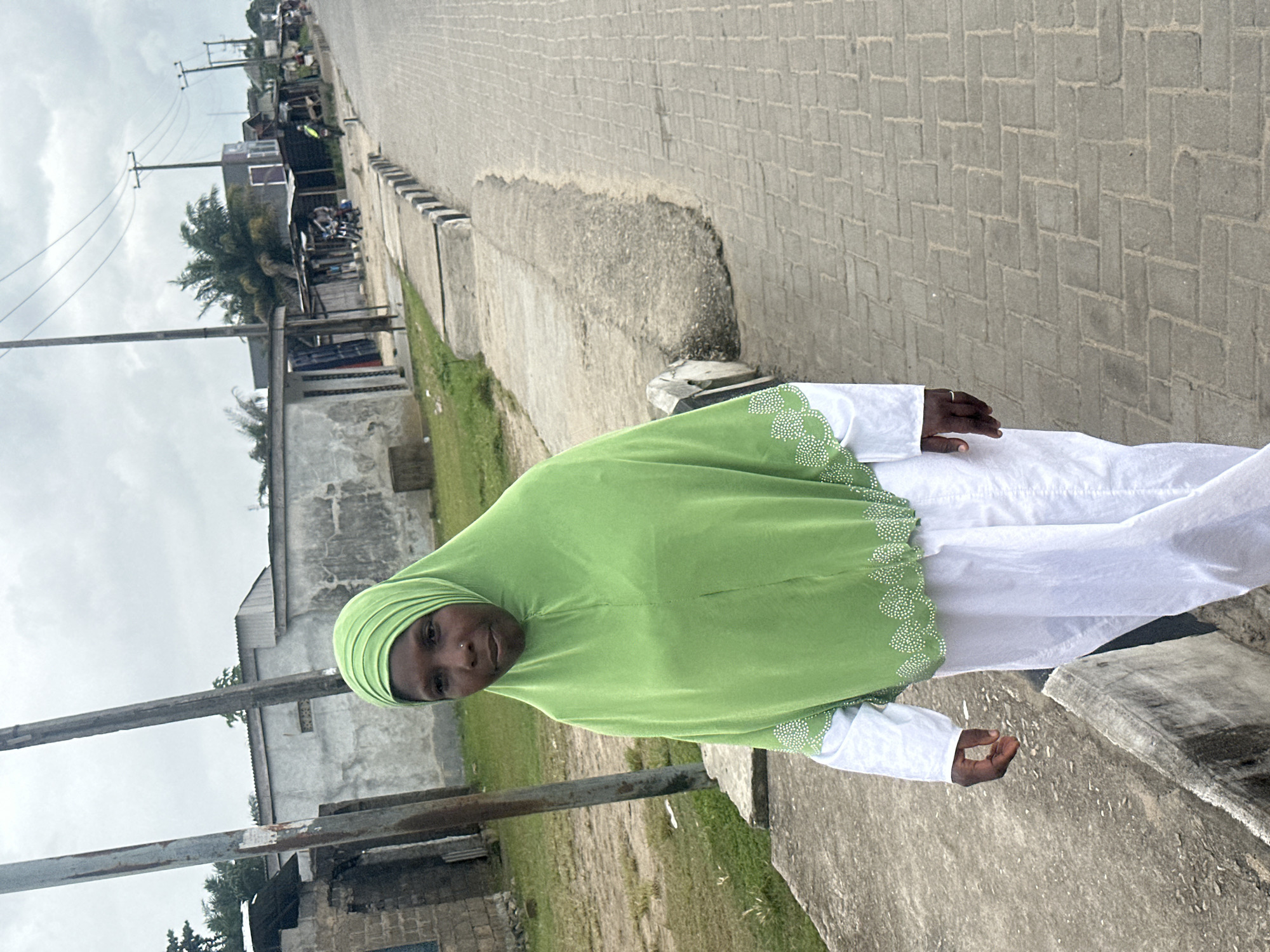 A woman wearing a bright green hijab and a white garment walks along a paved sidewalk in Badagry, Nigeria. She is positioned in the foreground, facing slightly away from the camera. Behind her, a street stretches into the distance, lined with buildings of various states of repair. Some buildings appear to be made of concrete with corrugated metal roofs, while others are more rudimentary structures with open fronts. Power lines crisscross the sky above. The environment suggests a typical urban or semi-urban setting in Nigeria. The paved sidewalk features a distinct pattern of interlocking bricks. The weather appears overcast.