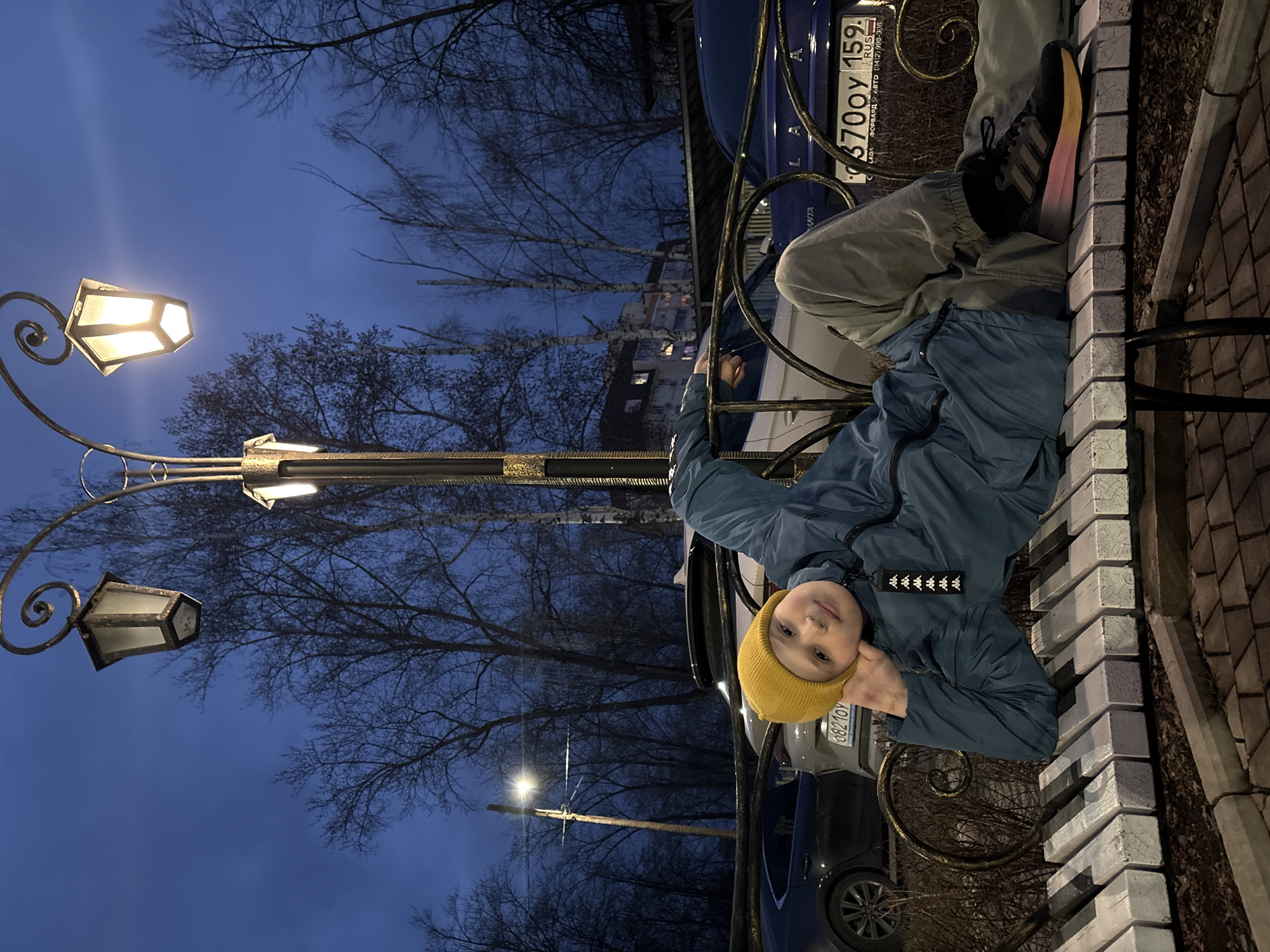 A young child is depicted sitting on an outdoor bench during the blue hour of evening or night in Chaikovsky, Russia. The child, wearing a bright yellow beanie, a blue padded jacket with a Kappa logo on the zipper pull, light gray pants, and black sneakers with a distinctive pink and yellow gradient sole, looks towards the camera with a calm expression, resting their chin on their hand.
The bench itself is a unique feature, designed to resemble a piano keyboard with alternating white and dark grey blocks forming the seating and backrest, supported by decorative dark metal scrollwork. The scene is illuminated by two ornate streetlights with warm yellow glows positioned in the foreground, and a brighter, simpler street lamp in the distance.
The background consists of bare, leafless trees silhouetted against a deep blue, twilight sky, suggesting a cold season. Faint outlines of distant buildings are visible through the trees. Several cars are parked behind the bench; a dark blue car displays a Russian license plate reading "370OY 159 RUS" and dealership details "LADA ФОРВАРД АВТО (3412) 998-308". Part of another license plate, "0821OY", is visible on a grey car. The ground adjacent to the bench is paved with cobblestone-like bricks.