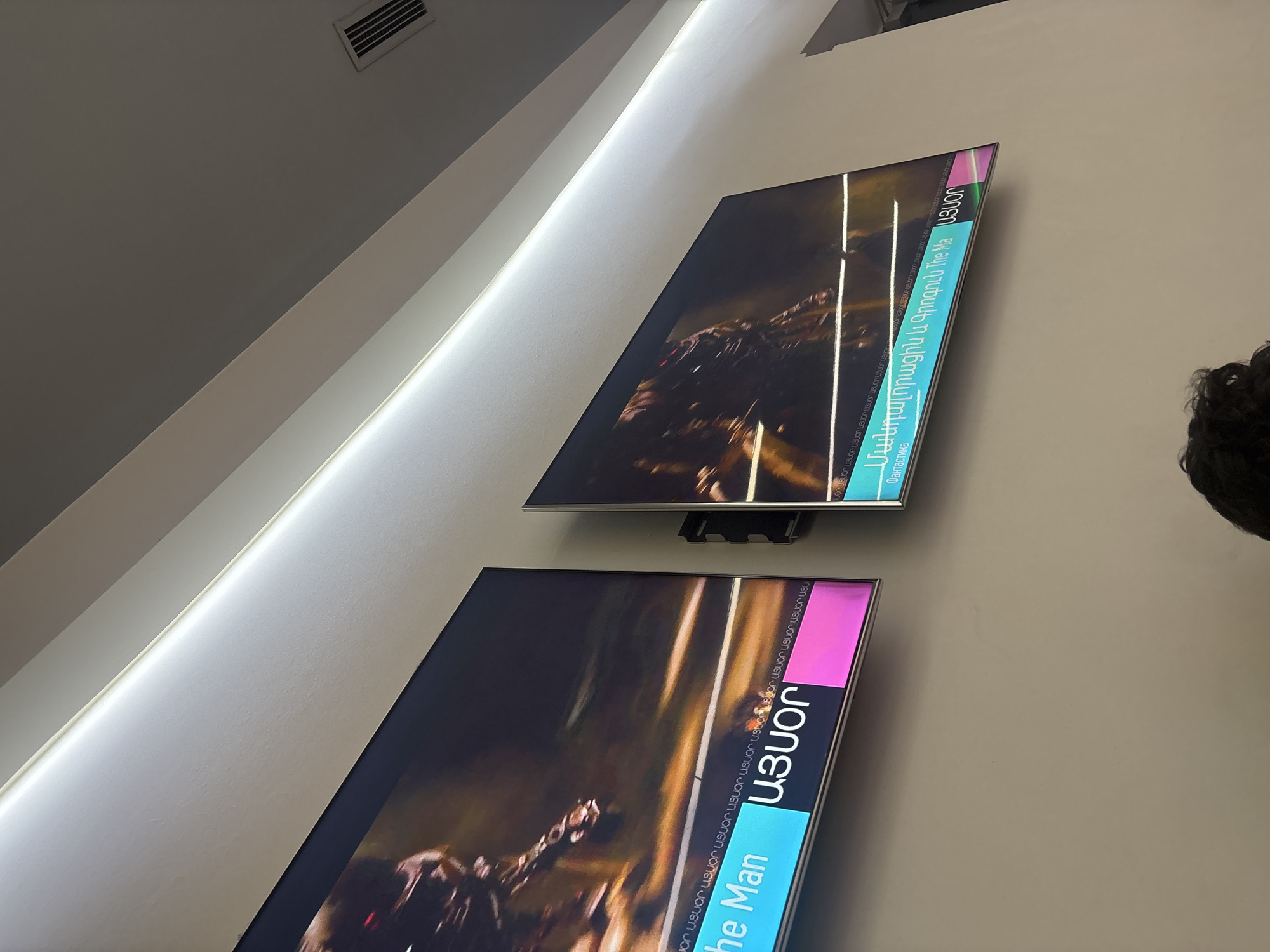 The image captures an indoor scene, likely a modern public space such as a cinema lobby or waiting area, with two flat-screen monitors mounted vertically on a light-colored wall. A linear light strip runs along the joint between the wall and the ceiling, providing illumination. An air vent is visible on the ceiling in the upper left corner.
The two monitors display promotional content, possibly for a movie or TV show. Both screens feature dark, action-oriented visuals, including what appears to be a creature or character. Prominently displayed text on the monitors includes "ԱՅՍՕՐ" (AYSO_R), which is Armenian for "TODAY." The upper screen clearly shows "Մանդալորացին և Գրոգուն" (Mandaloratsin yev Grogoon), translating to "The Mandalorian and Grogu," along with "Фантастика" (Fantastika), indicating the genre as Fantasy/Science Fiction. The lower screen also shows "The Man" and repeated instances of "ԱՅՍՕՐ" as a background pattern.
The photo is taken from a low angle, looking upwards at the screens. The top of a person's dark-haired head is visible on the right side of the frame, suggesting they are in the foreground, possibly observing the displays. The overall impression is a clean, contemporary setting with digital signage.