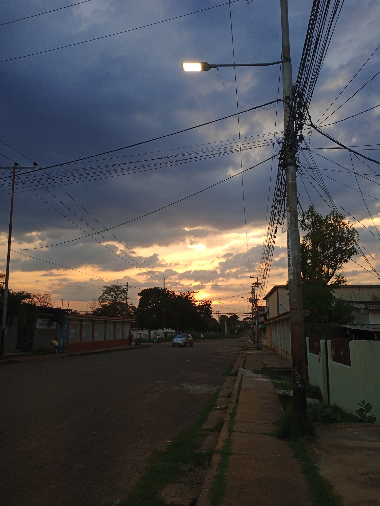 The image depicts a street in Valle de La Pascua, Venezuela, at dusk. The sky is partially obscured by dark clouds with an orange sunset visible in the distance. A street lamp is illuminated on a utility pole laden with numerous tangled wires. A few buildings are seen on both sides of the street, with varying architectural styles and conditions. A single car is visible further down the street. A few people are walking on the left side of the street. The road surface is cracked, and the sidewalk has patches of grass growing through the cracks. A partial advertisement "RUBEN ALCALA" can be seen on the pole.