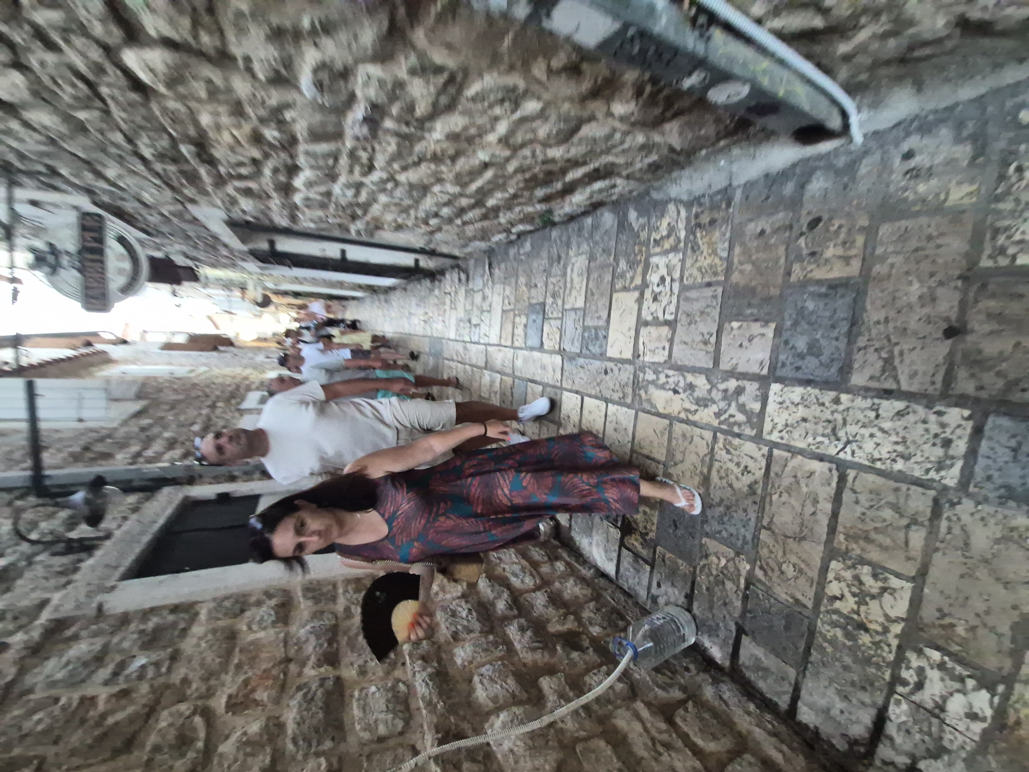 A diverse group of people are walking down a narrow, stone-paved alley in the Old Town of Budva, Montenegro. The walls are constructed of rough, aged stone blocks. In the foreground, a woman wearing a patterned dress and white flip-flops is walking away from the camera, holding a fan. To her right, a man in a white t-shirt and shorts follows. Further down the alley, more people, including children, are walking. A large plastic bottle filled with water and a hose are visible on the left side of the alley, near a stone wall. A sign with the word "RABBIT" is visible on the right wall. The lighting suggests it is daytime.