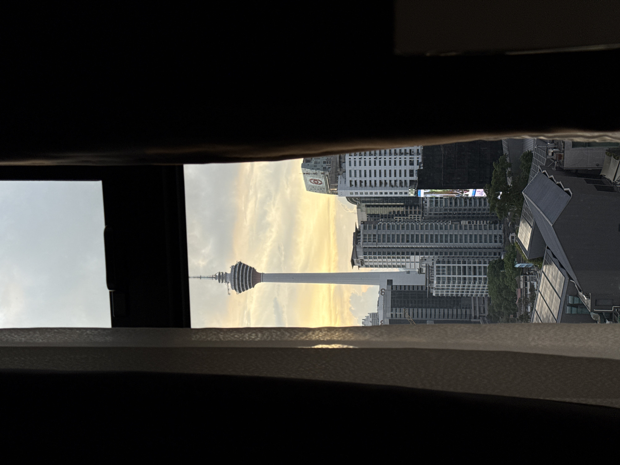 View of Kuala Lumpur Tower through a window. The tower is surrounded by other tall buildings. The sky is cloudy.