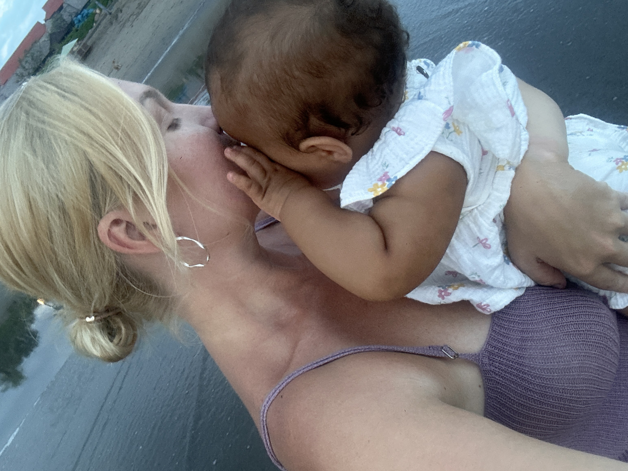 A woman with blonde hair tied in a bun is holding a baby close to her face on what appears to be a beach. The baby, dressed in a white floral patterned garment, has its hand near the woman's face. The woman is wearing a purple knit top and has silver hoop earrings. The background shows a shoreline with what looks like buildings or structures in the distance. The scene is possibly in North Kuta, Indonesia, given the coastal environment and probable location request.
