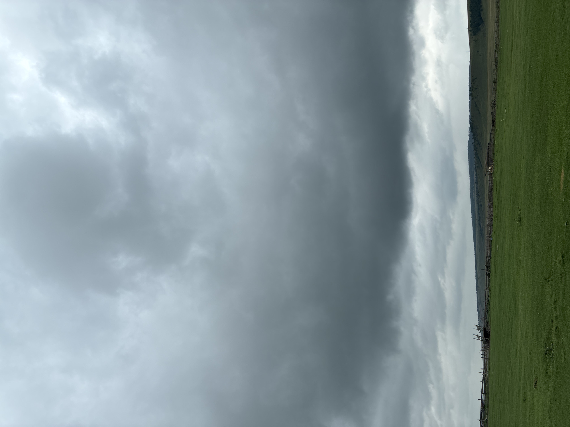 An exterior, medium shot captures a vast, open green field under a heavily overcast sky. The clouds are dark and dense, occupying the majority of the frame, suggesting an impending storm or recent rain. A wooden fence, appearing somewhat dilapidated and rustic, stretches horizontally across the middle ground of the image. Beyond the fence, a gently rolling landscape of hills and trees recedes into the distance, with more of the dark clouds visible above. The overall impression is one of a rural, possibly agricultural setting, with a somber, atmospheric mood due to the weather. The location is specified as Ust'-Ordynskii, Russia.