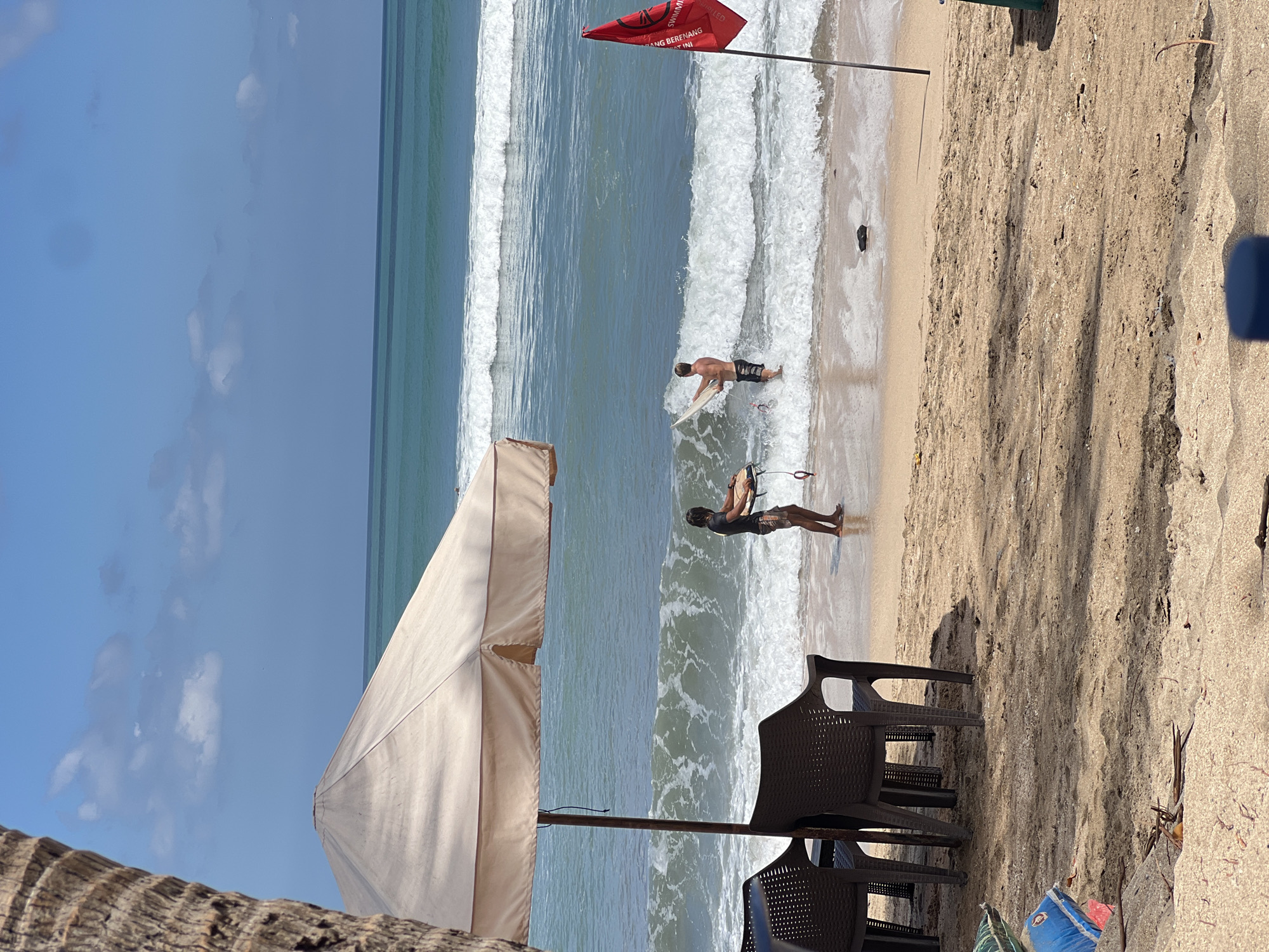 The image depicts a beach scene in Kuta, Indonesia, under a clear blue sky with scattered clouds. In the foreground, a large, tan beach umbrella shades two dark brown plastic chairs on the sandy beach. To the right of the umbrella, a red flag is planted in the sand, with text that appears to be in Indonesian. The ocean features turquoise water with rolling waves. Two individuals are visible in the shallow water, each holding a surfboard. One person, wearing dark shorts, is closer to the shore and appears to be standing with a surfboard. The other, in patterned shorts, is further out in the water, also with a surfboard. The sand on the beach is light brown and appears to have some debris scattered on it, including some plastic items and dry leaves. A portion of a palm tree trunk is visible on the far left edge of the frame, partially obscuring the view of the umbrella.