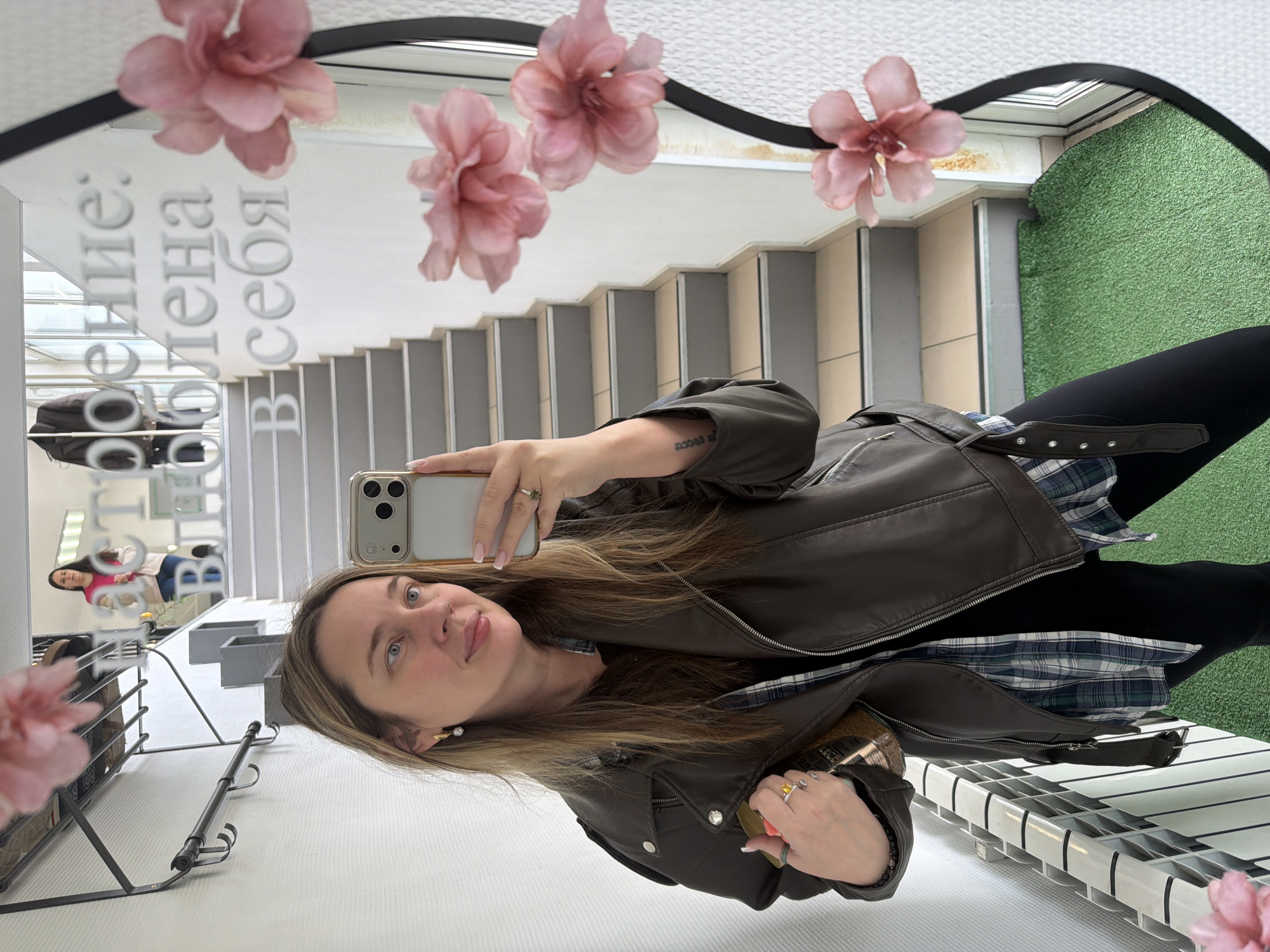 A woman takes a selfie in a mirror. The mirror is decorated with flowers. The reflection in the mirror shows a staircase, artificial grass, and a clothing rack. Another person is visible in the reflection as well. Text is visible in the mirror in Russian. The image was taken in the city of Chaikovsky, Russia.