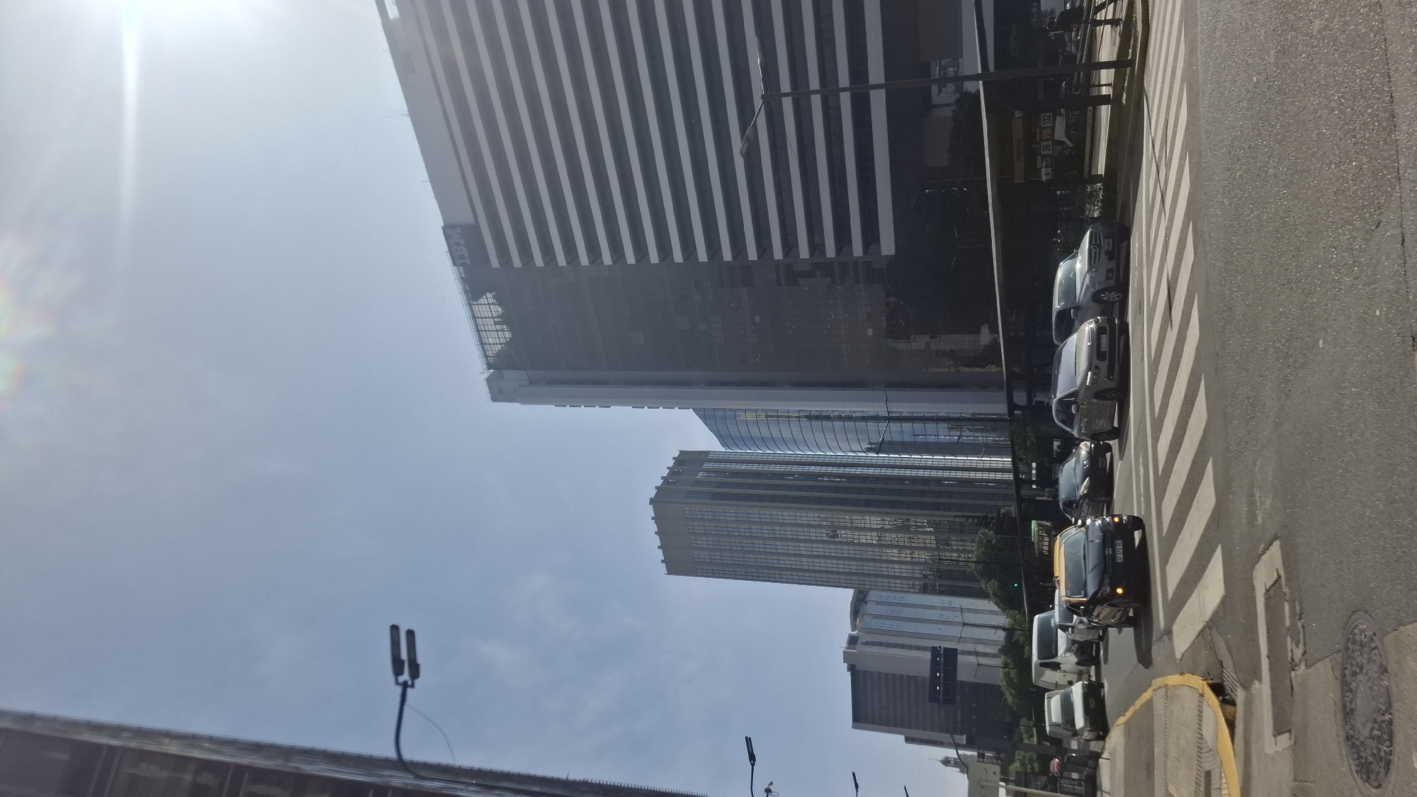 The image shows a street scene in Buenos Aires, Argentina, with a view of modern buildings and traffic. The prominent building on the upper left has a sign indicating it houses IBM. Several other skyscrapers are visible behind it. The street is lined with vehicles, including cars and at least one tuk-tuk or similar three-wheeled vehicle, stopped at a crosswalk. The image is taken from a low angle looking upward, against a bright sky, with possible sunlight glare. The scene is an urban environment, likely in the financial or business district.