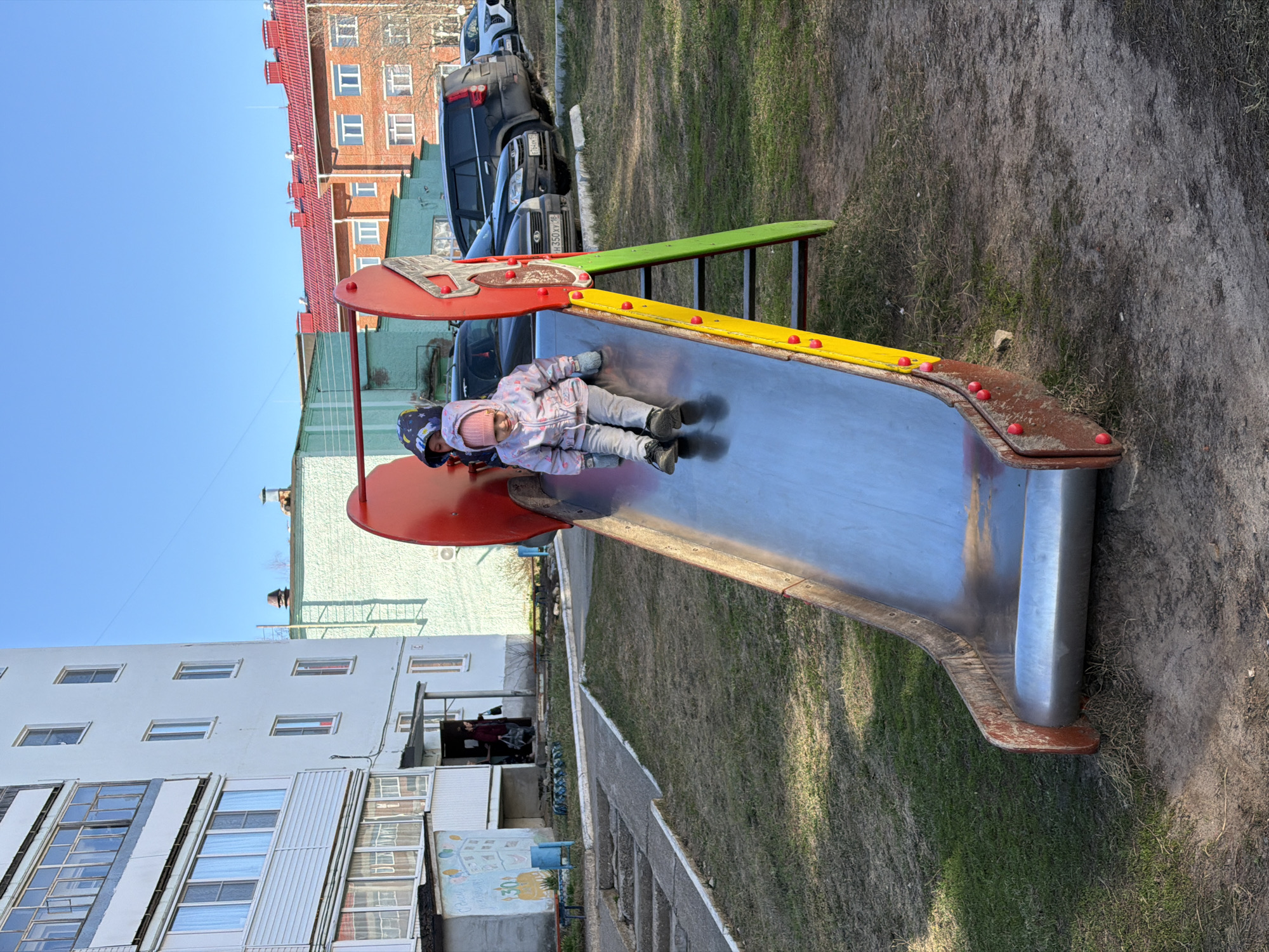 Two children are lying on their stomachs on a metal slide in an outdoor setting. The slide features a red, yellow, and green color scheme. Adjacent to the slide, multiple parked cars are visible. A building with red roofs and brick walls is located in the background behind the vehicles. The environment consists of dirt and grass. Another building with white walls and a row of windows is also present. The location is Noviy, Russia.