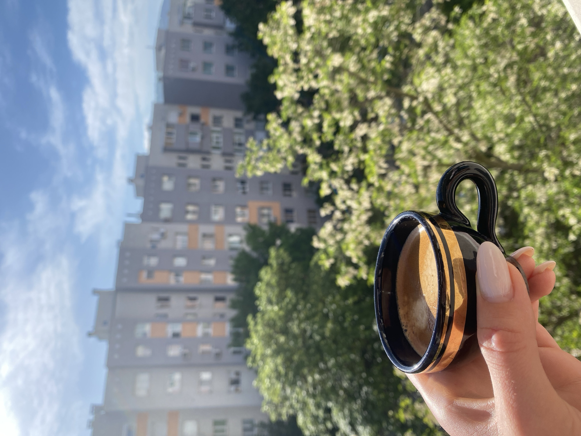 A hand holds a small cup of coffee in front of a building and tree in Blagoevgrad, Bulgaria.
The cup is black with gold stripes and filled with brown liquid. The hand holding the cup has pale skin and manicured nails.
Behind the cup, an out-of-focus view shows a multi-story building with a grey facade and a tree with green leaves. The sky is blue with white clouds visible above the building.