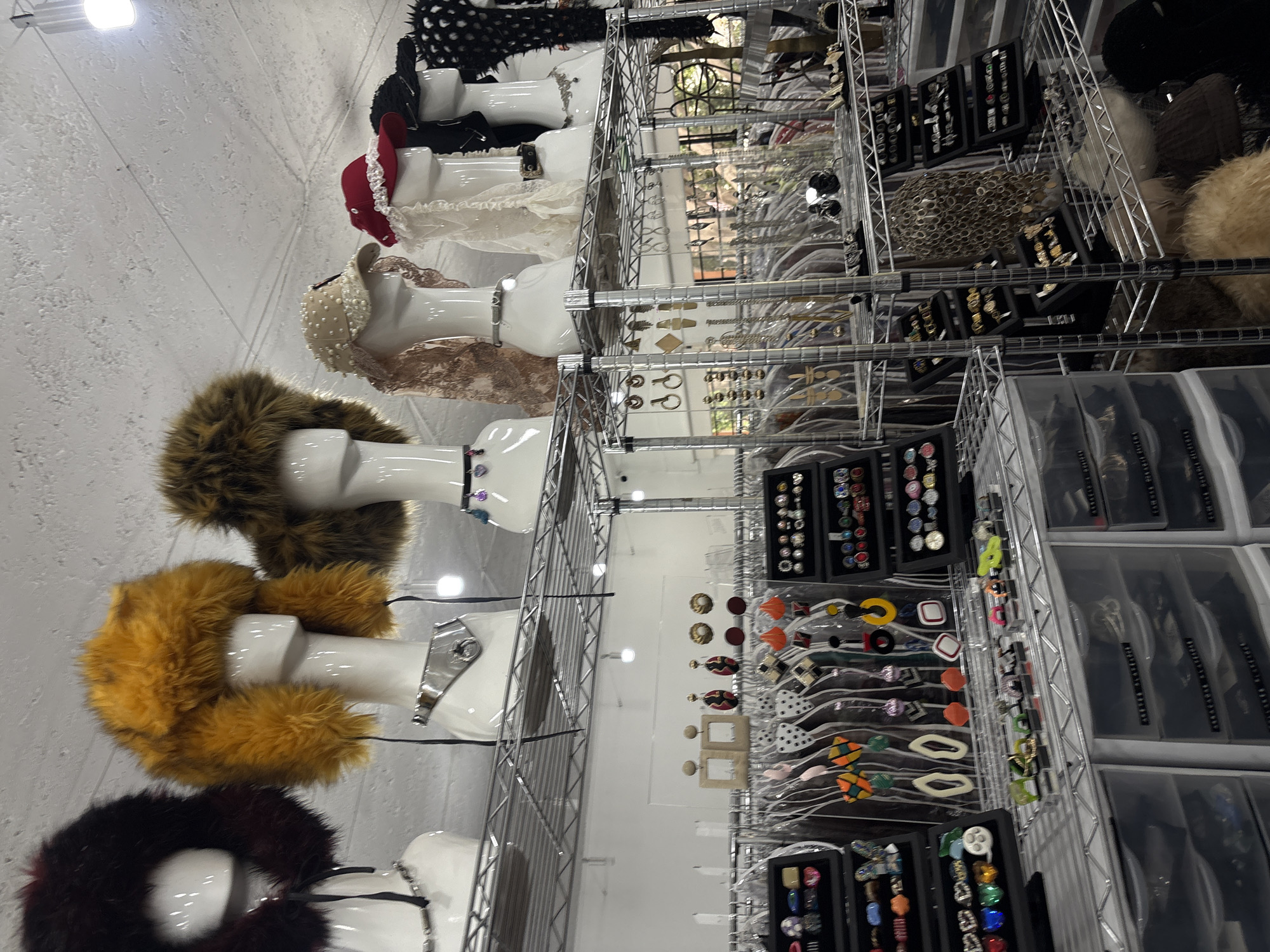 The image shows a display of fashion items on mannequins and shelves in what appears to be a retail environment. From top to bottom, the mannequins wear a variety of headwear, including a black spiked item, a red hat with white lace, a pearl-studded headpiece, a brown fur collar, a yellow fur collar, and a dark burgundy fur piece. The shelves next to the mannequins display jewelry, accessories, and organized collections of small items in clear containers. Given the context of the image, it is likely that this photo was taken in a store in Mexico City, Mexico, but there are no identifiable elements within the image that confirm the exact location.