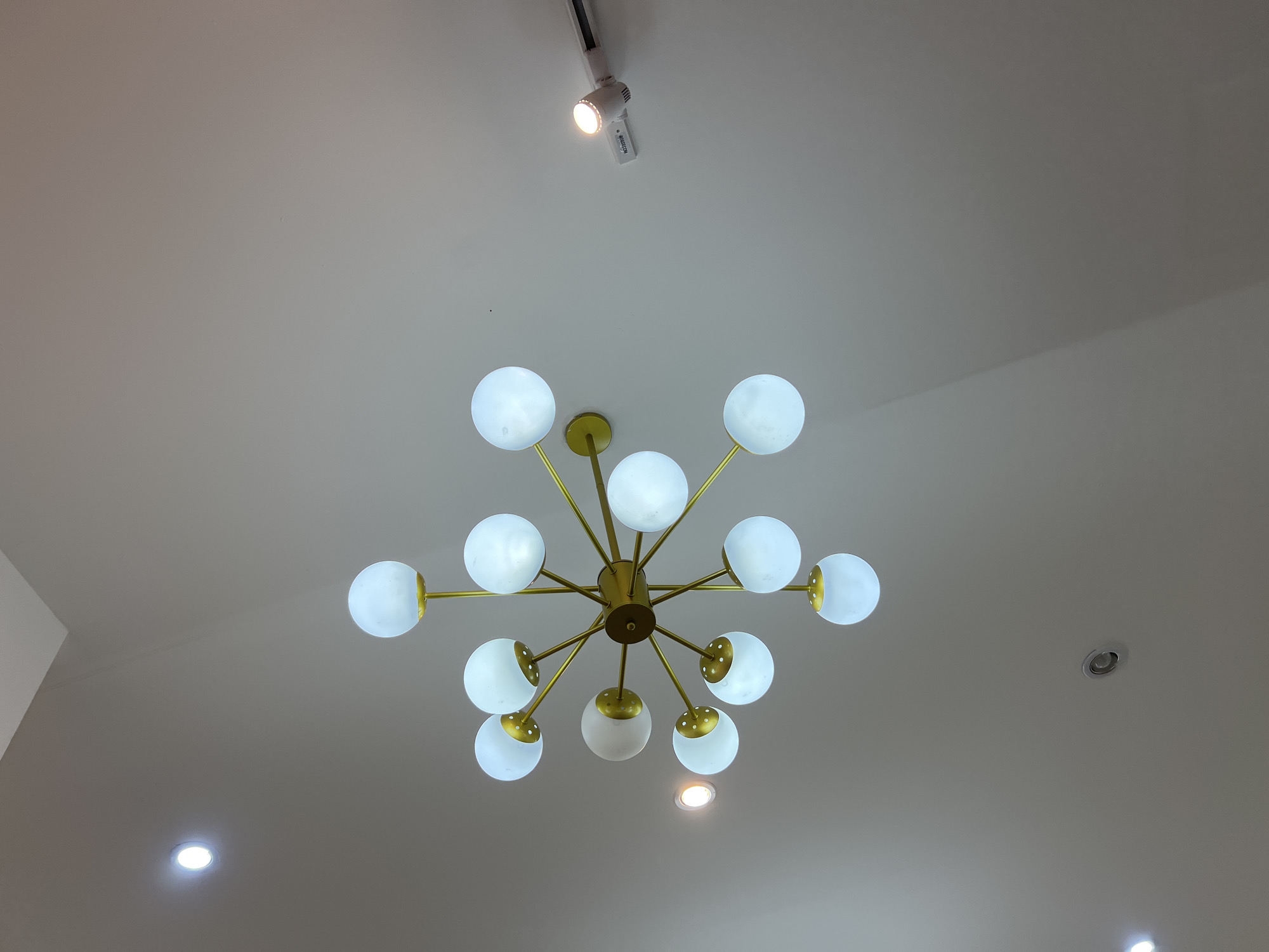 The image shows a modern, sputnik-style chandelier with multiple glowing white orbs attached to gold-colored arms, mounted on a white ceiling. There are also other light fixtures visible on the ceiling, including a track light with a bright bulb and a recessed ceiling light. The overall lighting is bright, suggesting an indoor setting. The image does not contain any information that would allow for the determination of its location within Denpasar, Indonesia, nor does it depict any people, actions, or interactions.