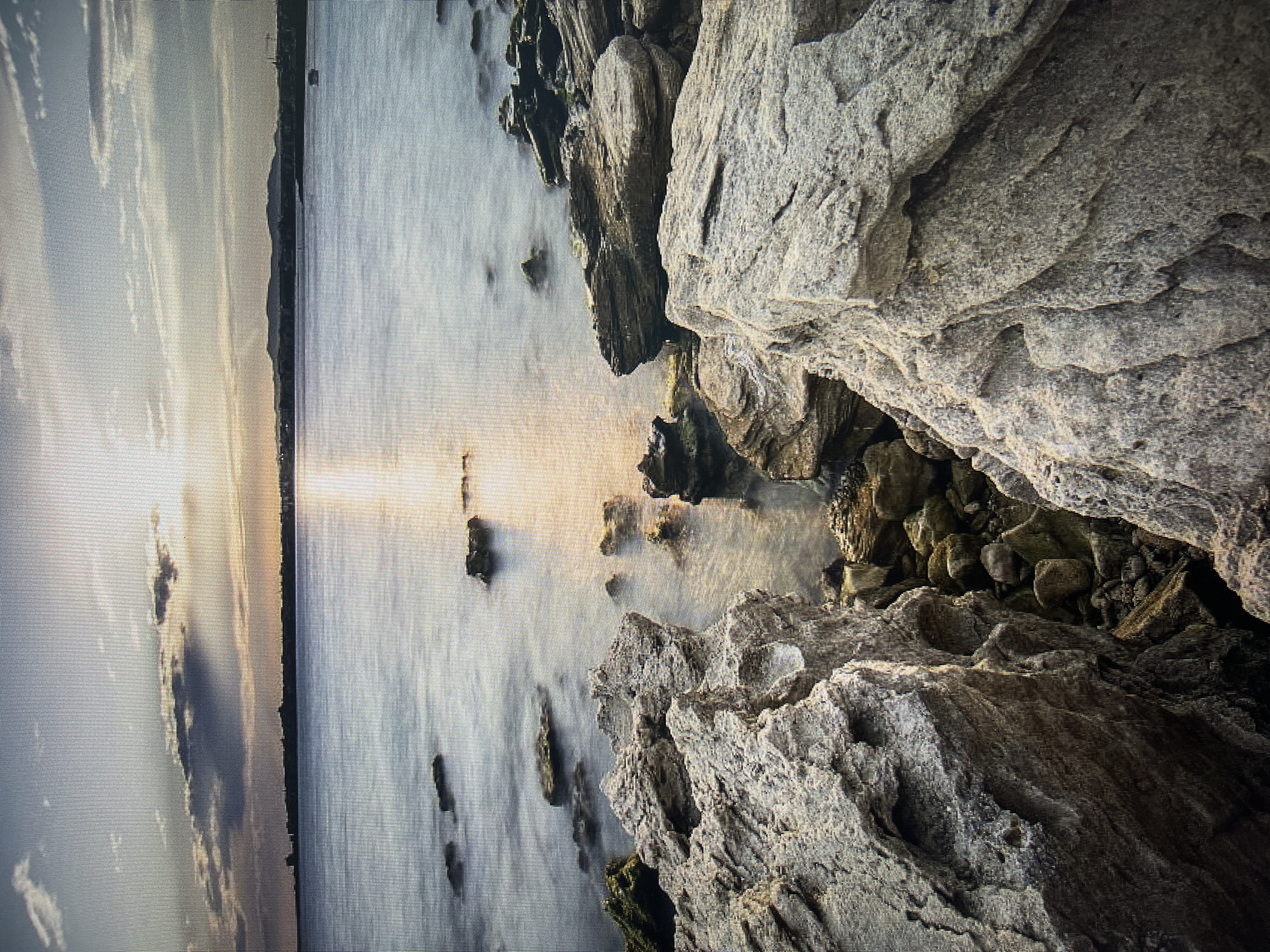 The image depicts a seascape with a rocky coastline in the foreground and the sea stretching towards the horizon. The sun is low in the sky, casting a warm glow and creating a reflection on the water's surface, suggesting either sunrise or sunset. There are several large, weathered rocks in the foreground, with smaller rocks and pebbles visible in the water. In the distance, a silhouetted shoreline with a town or city is visible, along with hills in the far background. The sky is partly cloudy, with sunlight breaking through.
The scene is consistent with the coastline of Varna, Bulgaria, specifically areas like the northern beaches or the rocky areas near the Varna Lighthouse, which offer similar views of the Black Sea with rocky outcrops and a distant city skyline. The long exposure used to capture the water's motion creates a smooth, almost ethereal effect on the sea. There are no people or discernible activities visible in the image, focusing solely on the natural landscape and the atmospheric lighting.