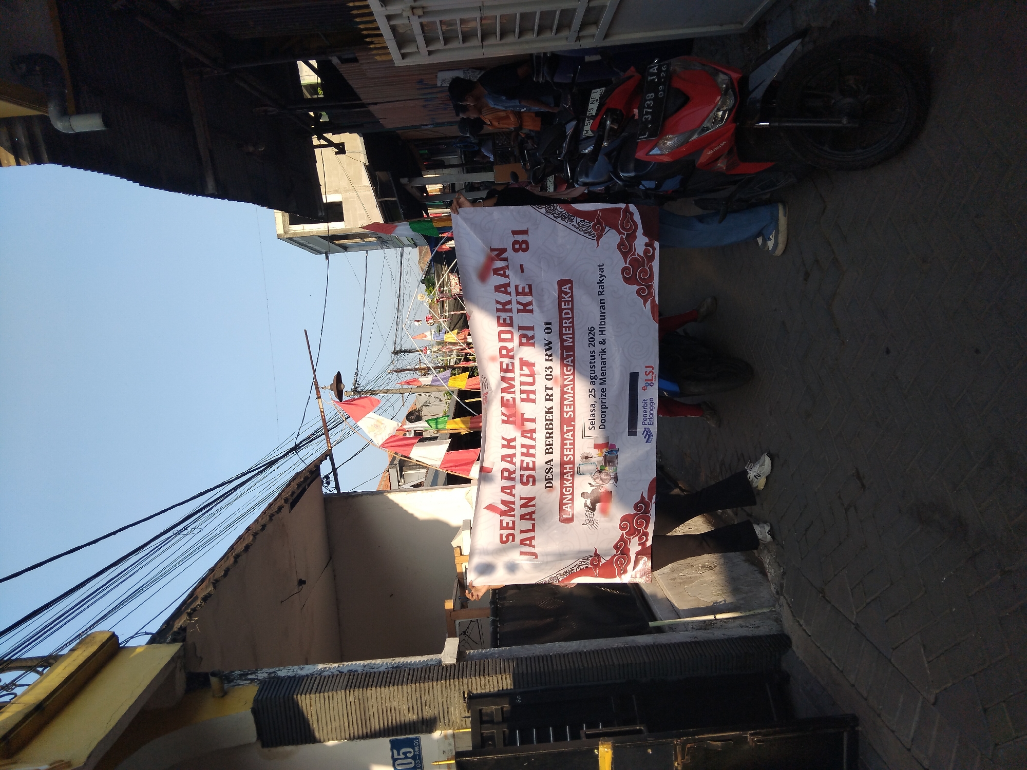 The image depicts an outdoor street scene in Kepuh Kiriman, Indonesia, during the day. A banner is held horizontally by at least three people, with their legs and lower torsos visible. The banner displays text in red and black, including "SEMARAK KEMERDEKAAN JALAN SEHAT HUT RI KE - 81" which translates to "Independence Celebration Healthy Road 81st Anniversary of the Republic of Indonesia". It also mentions "DESA BERBEK RT 03 RW 01", indicating the location as Berbek Village, RT 03, RW 01. Further details on the banner include "LANGKAH SEHAT, SEMANGAT MERDEKA" ("Healthy Steps, Spirit of Independence"), "Selasa, 25 agustus 2026" (Tuesday, August 25, 2026), and "Doorprize Menarik & Hiburan Rakyat" ("Interesting Doorprize & People's Entertainment"). Logos of "Penerbit Erlangga" and "BLSJ" are also visible.
Several motorcycles are parked in the street, including a red Honda motorcycle with license plate L 3738 JA. Flags, some red and white, are strung up in the background, suggesting a celebration. The environment appears to be a narrow street lined with buildings, some with a yellowish facade and tiled roofs. Overhead power lines are prominent. The overall lighting suggests it is daytime, with shadows cast on the brick-paved ground. Several people are visible in the background, some standing and observing.