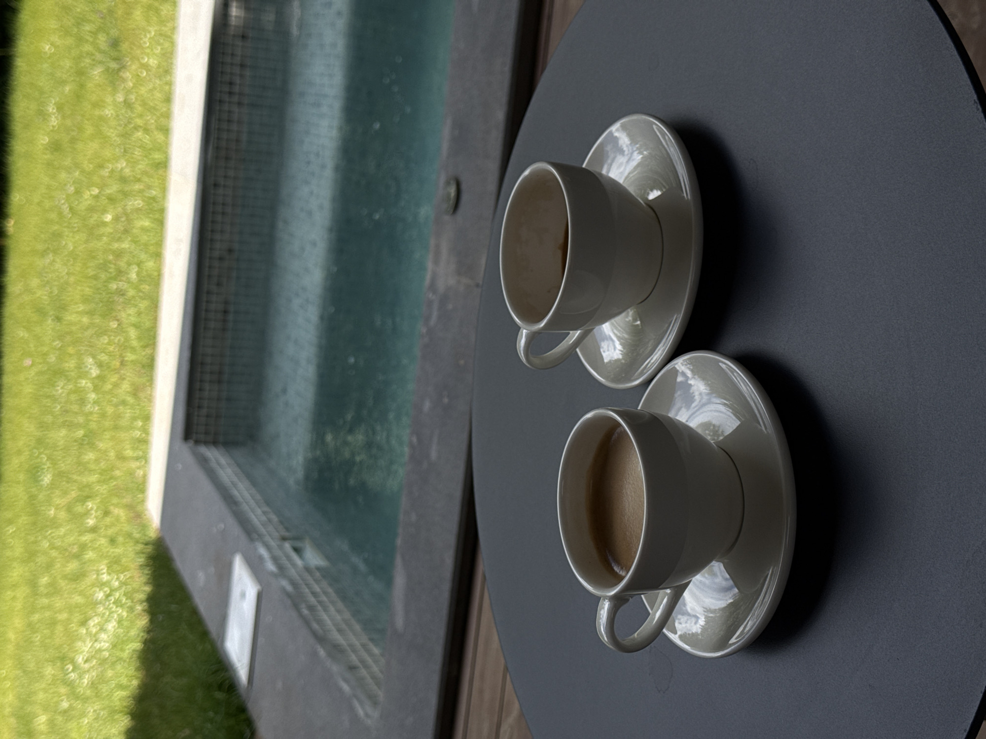 Two white ceramic coffee cups with saucers are placed on a round, dark gray table. One cup is filled with coffee. The table is positioned next to an outdoor pool area. To the left of the table, there is a section of bright green grass and the tiled edge of a swimming pool, which appears to be empty or very shallow. The water in the pool has a greenish-blue hue. The scene suggests a peaceful setting, possibly during the daytime, with the sunlight reflecting on the pool water and the glossy surfaces of the cups and saucers. The overall impression is one of relaxation and leisure.