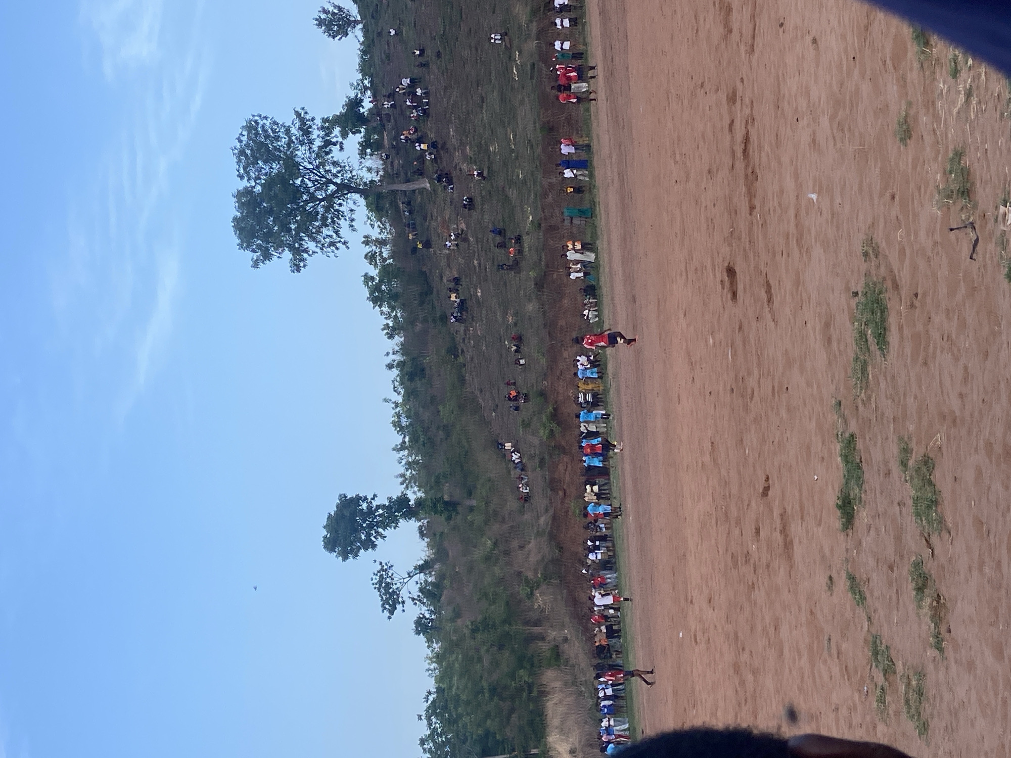 The image depicts a scene outdoors, likely in a rural or semi-rural area, possibly near Lokoja, Nigeria. A large dirt field occupies the right side of the frame, showing some sparse vegetation. A group of people, possibly a crowd or audience, lines the edge of the field, some sitting on a hill or slope behind the line of standing people. Their clothing suggests a mix of colors and patterns. A tree stands near the field. The sky is visible. An individual stands on the field.
