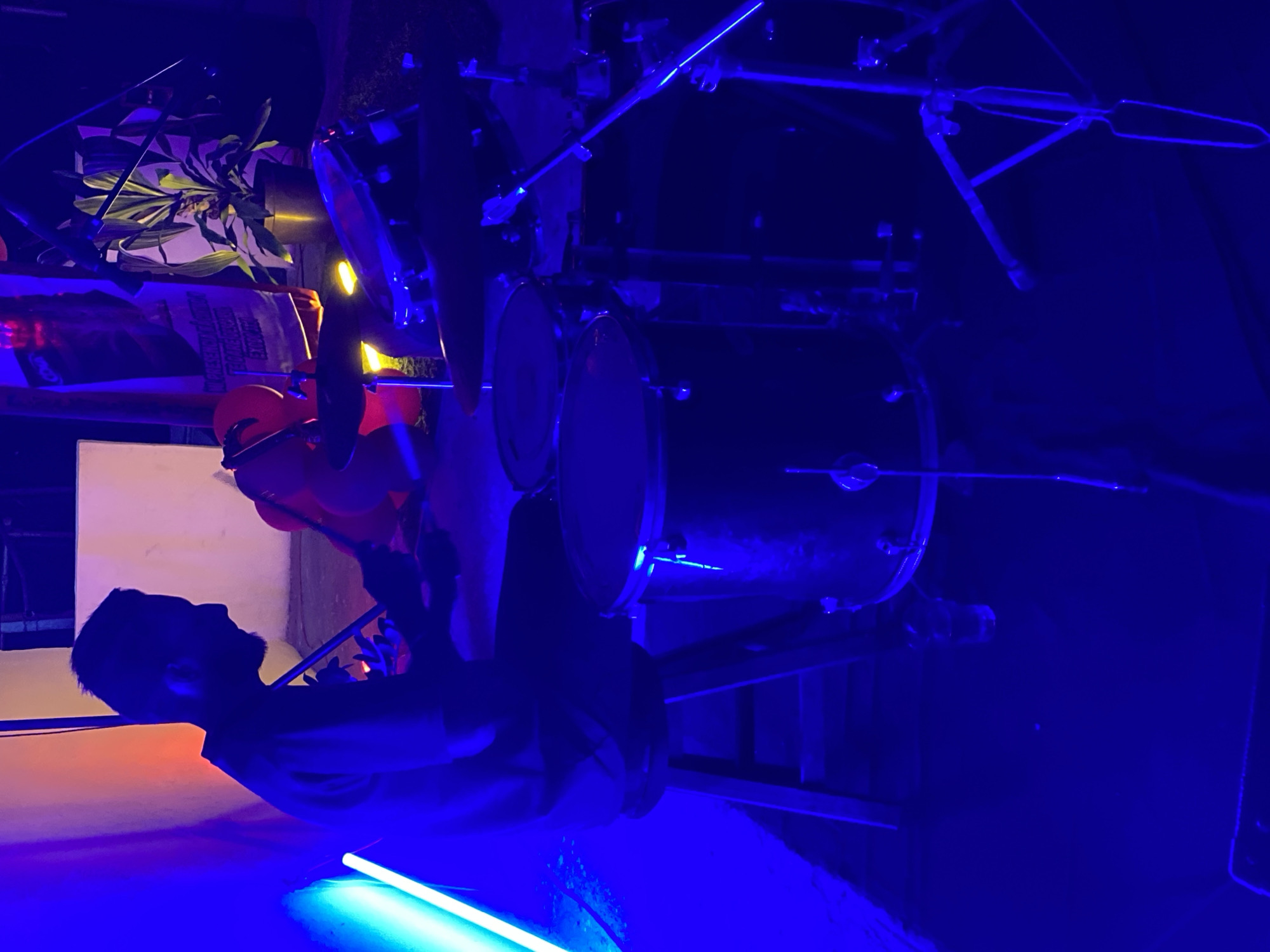 A silhouetted figure of a man, viewed from the side, is standing next to a drum set under blue and orange lighting. The location is Tinaquillo, Venezuela.