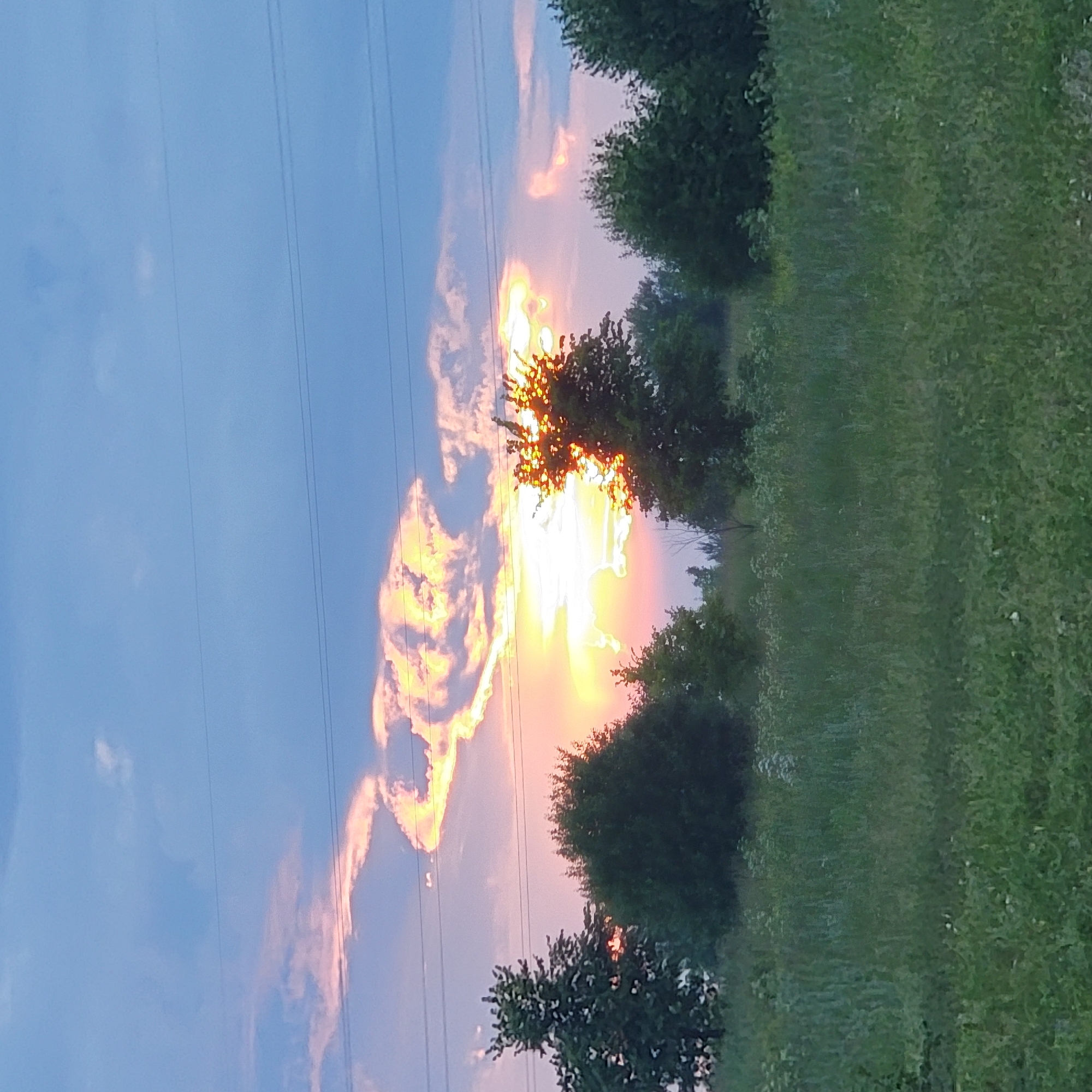The media file depicts an outdoor scene with a sunset. The sky is a mix of blue and vibrant orange and yellow hues, indicative of the sun setting. Several clouds are illuminated by the setting sun, creating a dramatic effect. Thin, dark power lines stretch across the sky from left to right.
On the right side of the frame, a grassy field slopes upwards, lined with several trees that are silhouetted against the bright sky. The trees are dark green, and their foliage appears dense. The grass is also green, with some areas appearing lighter, possibly due to sunlight. The overall setting suggests a rural or semi-rural location.
The time of day is clearly sunset, given the colors in the sky and the lighting. There are no people or specific objects visible, other than the natural elements of the landscape and the man-made power lines. The mood conveyed by the image is one of natural beauty and tranquility, with the dramatic sunset adding a sense of awe. No text is visible in the image. The location context is Ufa, Russia.