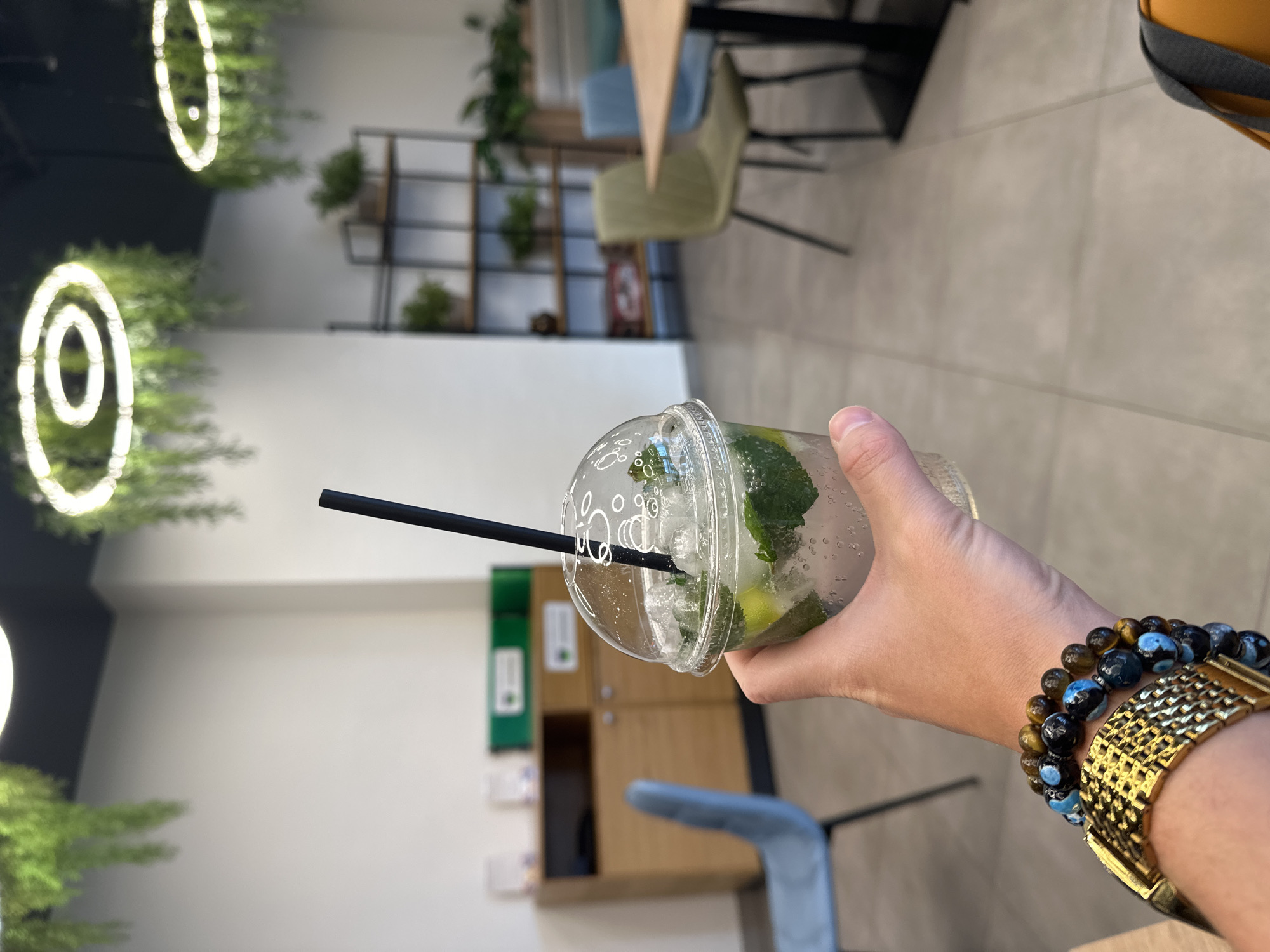 A hand holds a clear plastic cup filled with ice, mint, and a dark straw. The foreground shows a wrist adorned with a gold watch and two beaded bracelets, one blue and black, the other gold and brown. The background includes a neutral-toned wall and floor, chairs, and what appears to be a shelving unit with plants. Round light fixtures adorn the ceiling.
The setting appears to be indoors in a modern space. There is no indication the image was taken in Moscow, Russia.