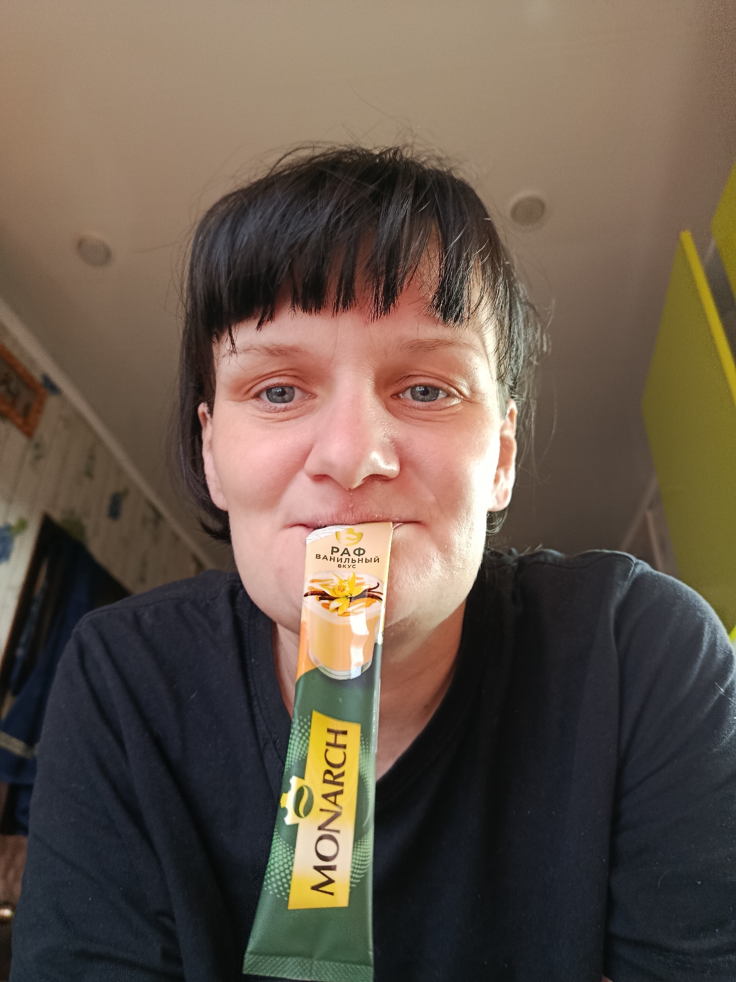 A woman with short, dark hair and bangs smiles slightly at the camera, holding a green and yellow sachet of an instant drink mix to her mouth. She has light blue eyes and is wearing a black t-shirt. The sachet is branded "MONARCH" in yellow text, with a small coffee bean logo. Above this, the packaging features an image of a drink in a glass with vanilla beans and a flower. Cyrillic text on the sachet reads "РАФ ВАНИЛЬНЫЙ ВКУС," translating to "RAF VANILLA FLAVOR."
The scene is indoors, likely a home in Фоки, Russia. The background on the left features patterned wallpaper, possibly with a blue floral design, and a dark curtain or fabric. On the right, a bright yellow-green surface is visible. The ceiling is light-colored and seen above the woman's head. The lighting is soft and even, suggesting daytime. The woman's expression is pleasant and relaxed, indicating a casual moment of enjoying her drink.