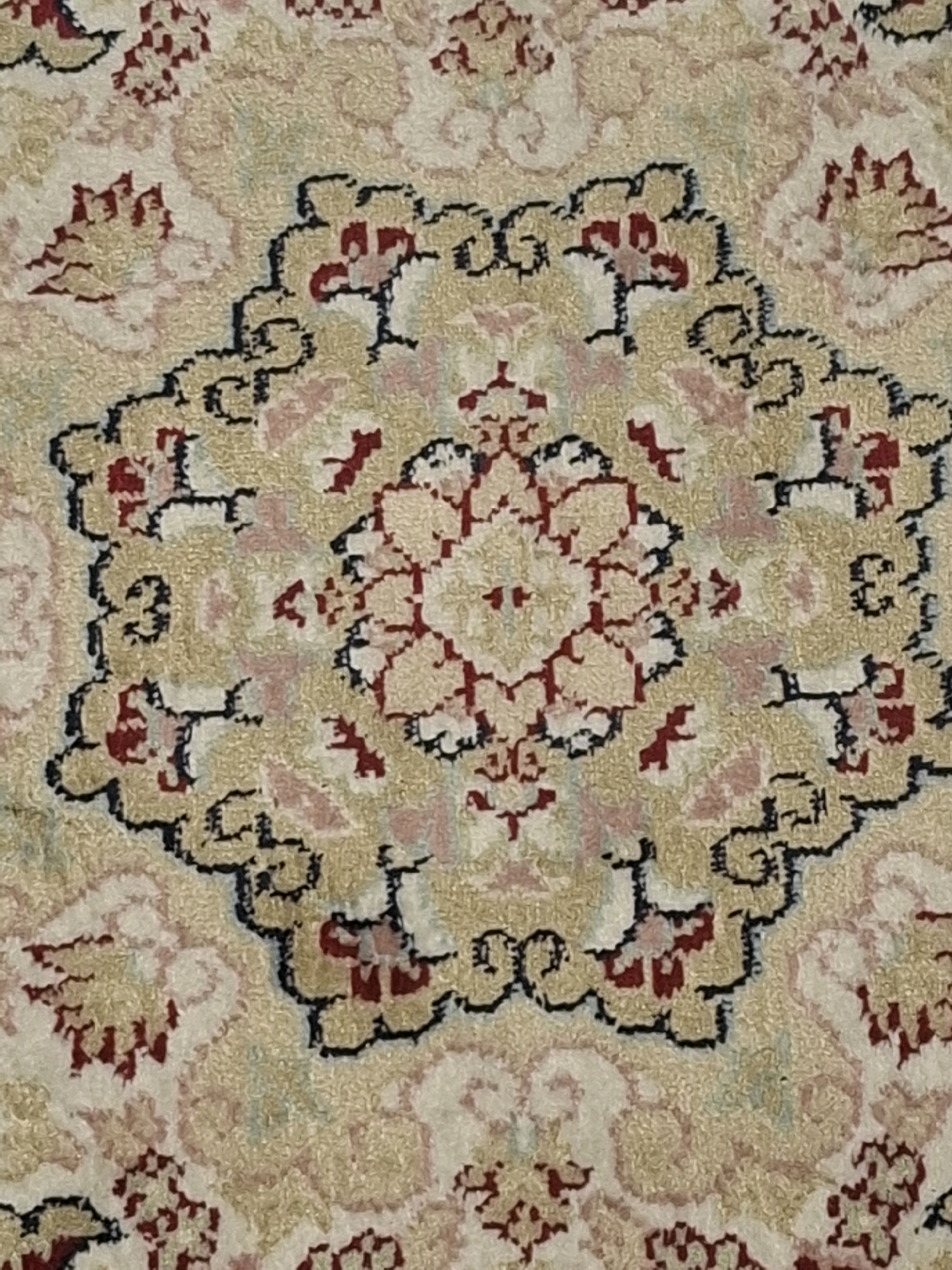 The image shows a close-up, overhead view of a decorative carpet with a central medallion design. The carpet features a complex geometric and floral pattern in shades of cream, beige, red, and black. The central medallion is an intricate eight-pointed star shape with floral motifs and scalloped edges. This medallion is surrounded by further decorative elements, including floral sprays and curved, almost abstract shapes, all outlined in black. The texture of the carpet appears to be plush and woven. The image does not provide any context or elements that would allow for the identification of a specific location in Almaty, Kazakhstan.