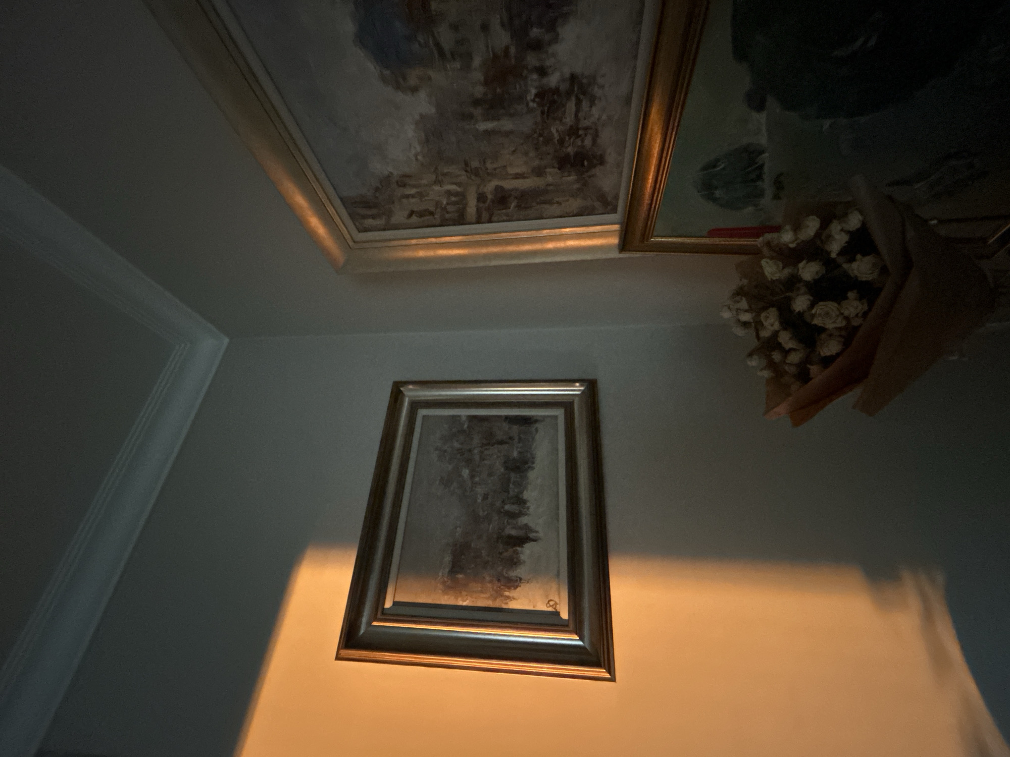The image depicts an indoor corner of a room, likely a home, with two framed paintings on the wall. A warm, orange light casts a sharp shadow across the wall and the left painting, suggesting a light source from the right. The painting on the left is horizontal, featuring a scene with figures in what appears to be a snowy landscape. The painting on the right is larger and vertical, depicting an urban scene with buildings. In the foreground, a bouquet of dried white flowers is arranged in a brown paper wrapping. The room has white crown molding along the ceiling. There are no people visible in the image. The scene is static and quiet, with the light and shadow creating a dramatic effect on the artwork. This scene is not identifiable as being in Plovdiv, Bulgaria, as there are no specific architectural or environmental cues present that would indicate its location.