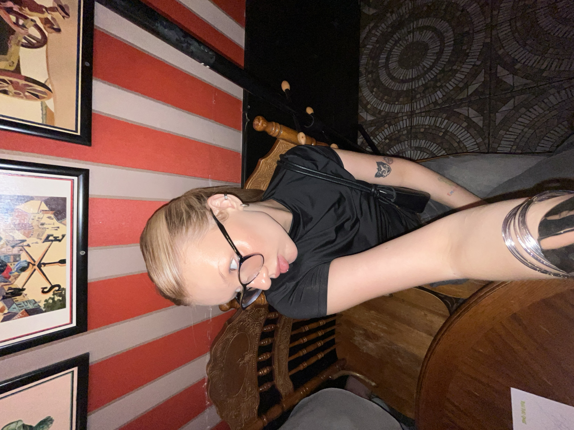 A light-skinned person with blonde hair and glasses is indoors at a restaurant in Chaikovsky, Russia, as suggested by the menu. The person is sitting on a wooden chair, wearing a black shirt, with multiple bracelets on their right wrist and tattoos on their left arm. The restaurant has red and white striped walls with framed pictures. The lighting is dim.