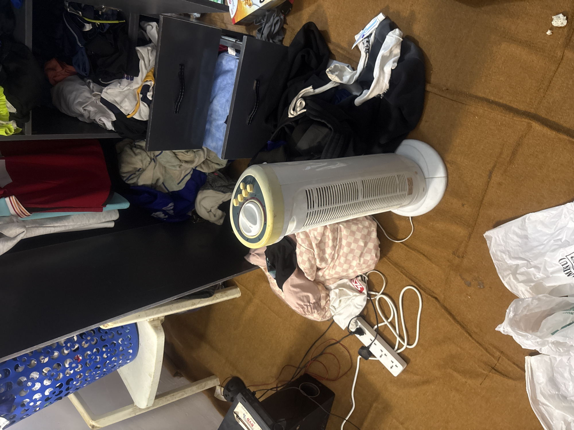 The image depicts a cluttered interior scene. In the foreground, a white tower fan lies horizontally amidst piles of clothing, including a pink checkered garment. A power strip with multiple plugs is visible, connected to various electronic devices. A black storage unit with open drawers, overflowing with clothes, is present on the left side. A blue laundry basket sits below the unit. In the background, a brown carpet or rug covers the floor, partially obscured by plastic bags. The location is identified as Gura Top, Nigeria. No people are visible.