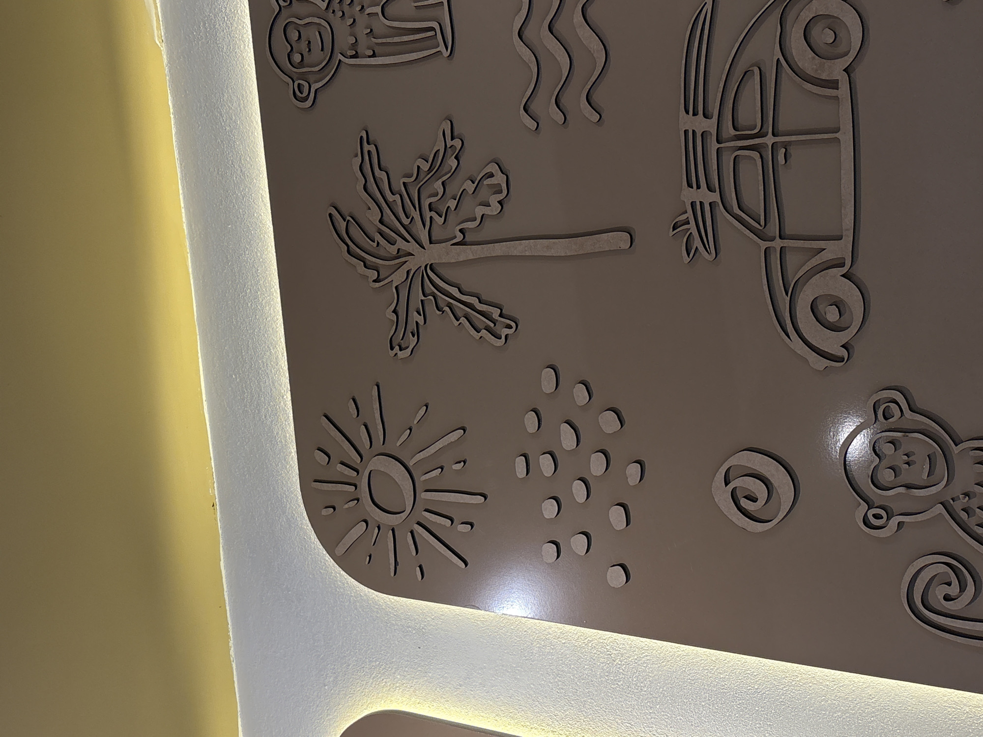A close-up shot reveals a section of wall art featuring laser-cut or routed designs in a tan or light brown material, possibly wood or a composite. The designs include recognizable shapes such as a monkey, a palm tree, a sun, a car, wavy lines, and abstract shapes. The wall art is adjacent to a surface that appears to be painted a light yellow. A section of a textured white surface, likely part of the building's architecture, runs vertically along the left side of the wall art and features inset lighting that casts a warm glow. The location is not discernible from the image.