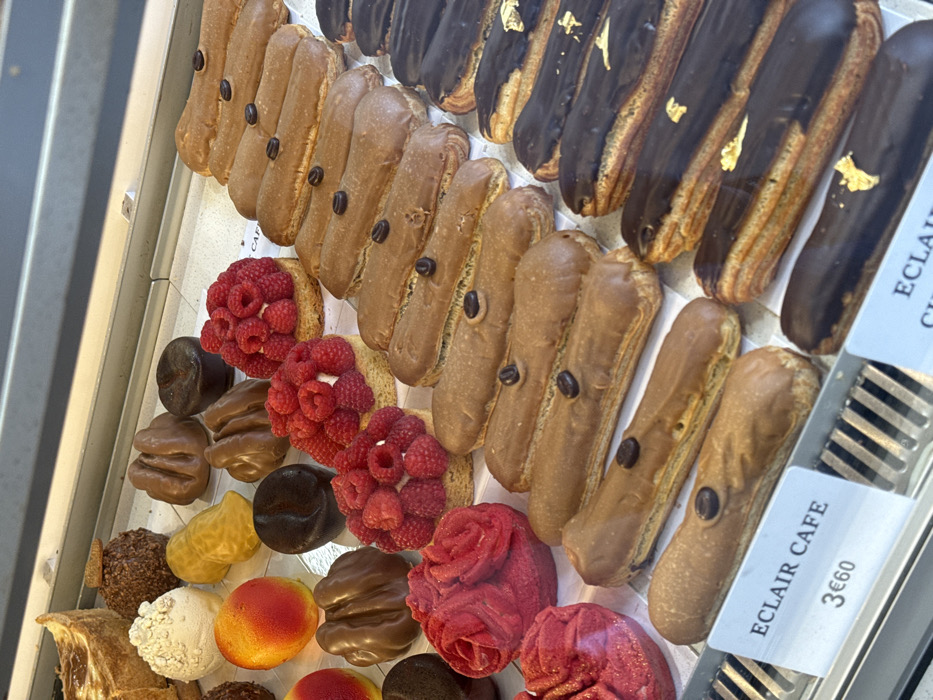 The image depicts an interior view of a pastry shop display case in Paris, France. An assortment of meticulously arranged French pastries and desserts fills the glass-fronted, refrigerated unit, resting on a white surface.
The most prominent subjects are multiple rows of eclairs. On the right, several rows of coffee eclairs are visible, each featuring a light brown frosting and individually topped with a single dark coffee bean. Below these are rows of dark chocolate eclairs, distinguished by their rich, dark frosting adorned with small, irregular flakes of gold leaf. A white price tag in the foreground identifies an item as "ECLAIR CAFE" and lists the price as "3€60."
To the left of the eclairs, various individual pastries are arranged. These include several round tartlets with a shortbread base, filled with cream and topped abundantly with fresh red raspberries. Two distinct, deep red, rose-shaped pastries are present, crafted with intricate petal details. Additionally, there are multiple brown, irregularly shaped pastries; some are dark brown and somewhat mushroom-like, while others are lighter brown and appear ridged. Two bright, spherical pastries with a red-to-yellow gradient, resembling peaches or apricots, are also displayed. A single white, textured, spherical pastry and a larger, partially visible tart-like pastry are located in the upper left section of the case.
The environment indicates a retail setting, with no individuals visible within the frame. The display case features a reflective interior and metal grates along the bottom edge.