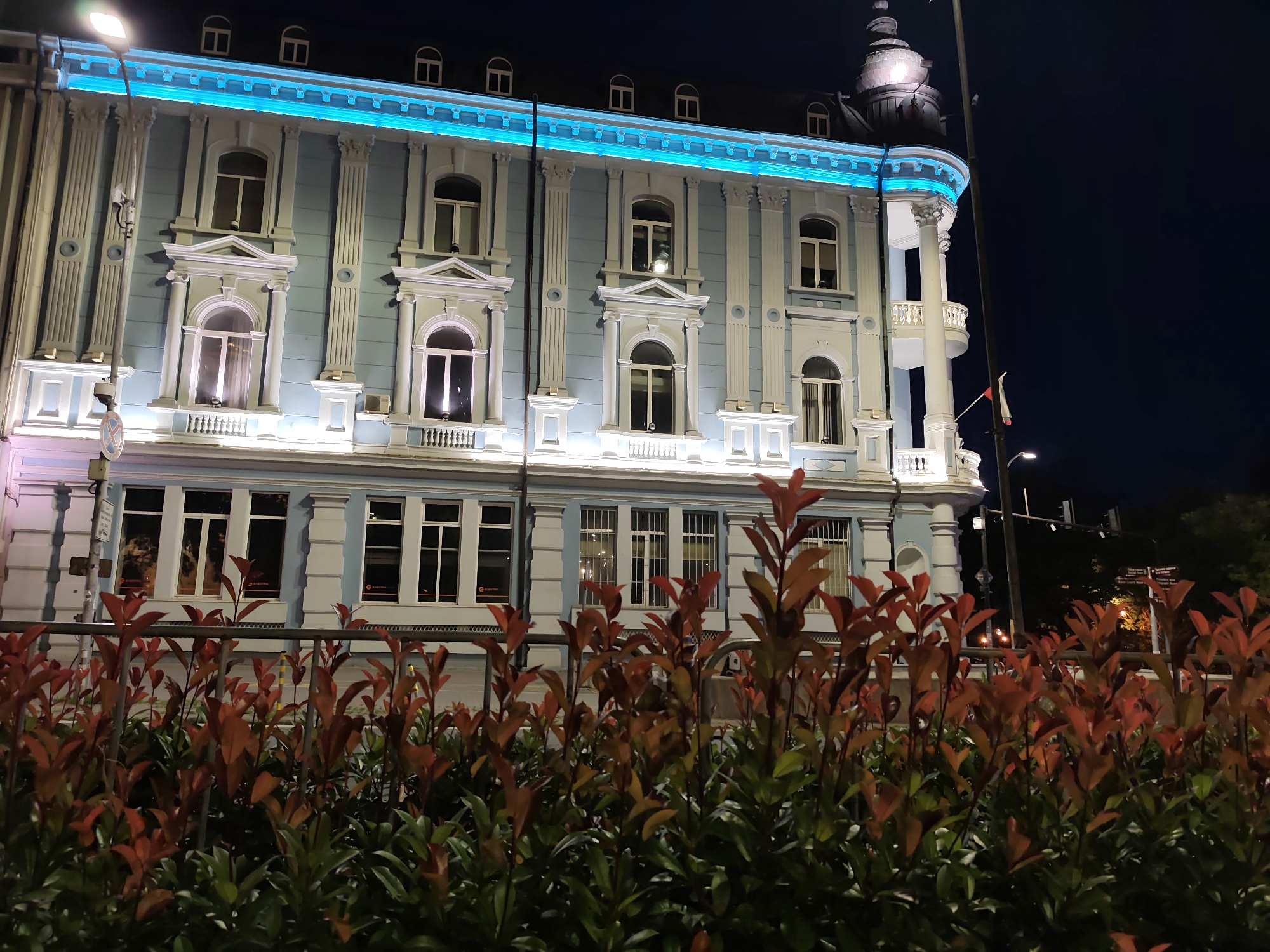 The image depicts a building in Varna, Bulgaria at night. The building is illuminated and features classical architectural elements, including columns and arched windows. A bright blue light runs along the top of the building. In the foreground, there is a dense hedge with red and green leaves, partially obscuring the lower part of the building and a metal fence. A street lamp is visible on the left side of the frame, and a flag on a pole is to the right of the building's corner. The overall scene suggests an urban environment at night, with the building being a prominent feature.