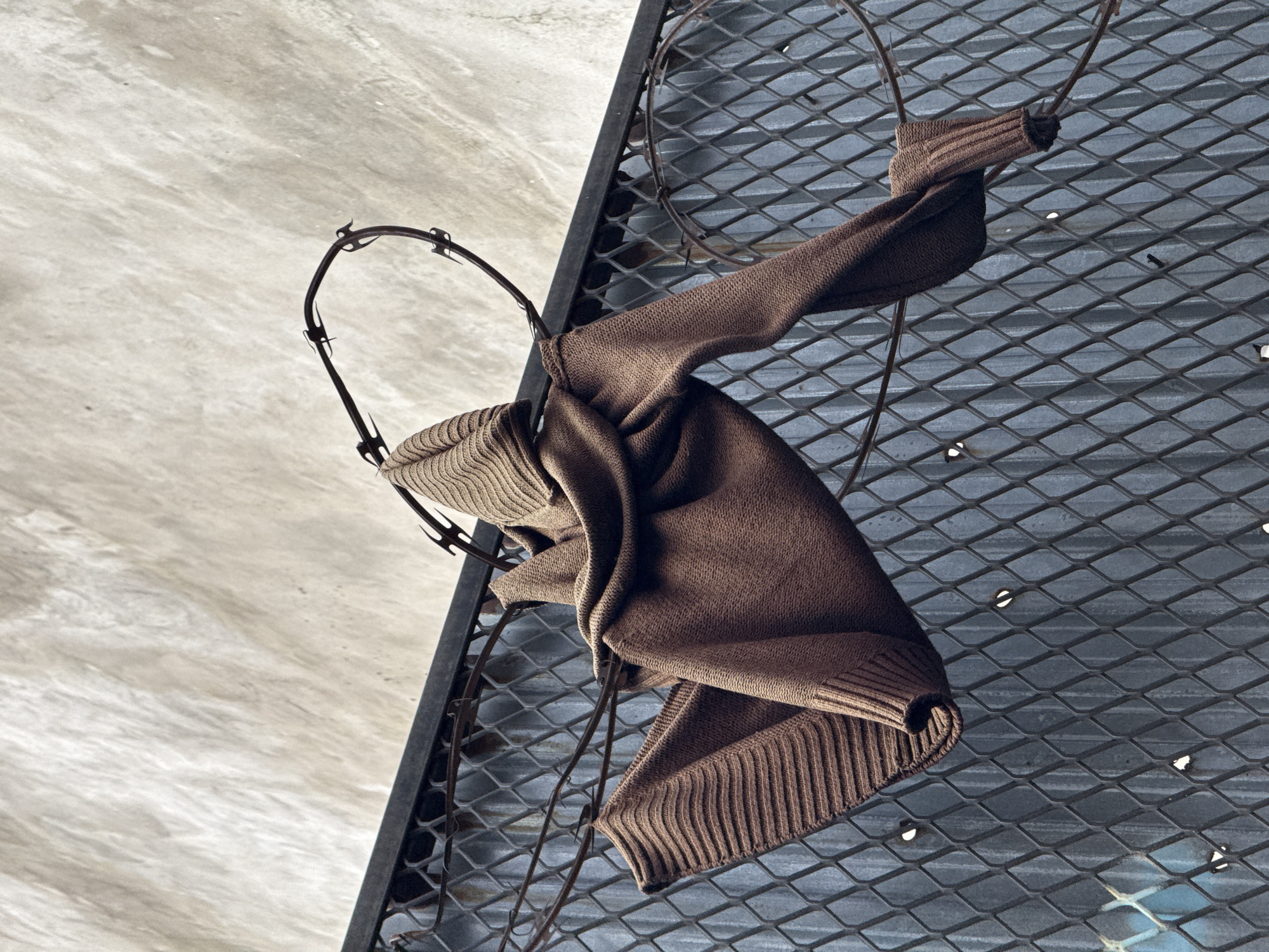 A brown knit garment, consistent with a sweater or long-sleeved shirt, is draped over and partially impaled by strands of rusted barbed wire. The barbed wire is positioned along the top edge of a dark gray, diamond-patterned metal fence or grating, which features several small, reflective points. One section of the barbed wire forms an irregular, incomplete coil. Behind the metal fence, a blue-gray corrugated metal surface is visible. Above the fence and barbed wire, a light-colored, textured wall, consistent with concrete or stucco, occupies the upper portion of the frame. No individuals are present. The scene is located in the city of New York, United States of America.