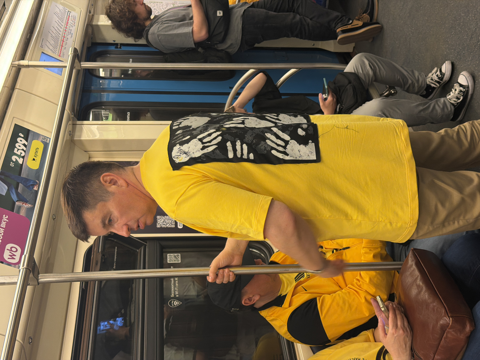 This indoor shot depicts a crowded scene inside what appears to be a subway car. A fair-skinned man in a yellow shirt and khaki pants holds onto a steel grab bar. The shirt has a black patch on the back with white handprints. A person next to him wears a yellow jacket with black accents and uses a cell phone, with a brown bag nearby. Other passengers include a person in a gray shirt and black pants, lying on the upper seating area, and a seated passenger wearing gray pants and black sneakers with white laces. The subway car has light beige walls with advertisements and information panels, as well as dark doors. A blue window panel is visible.