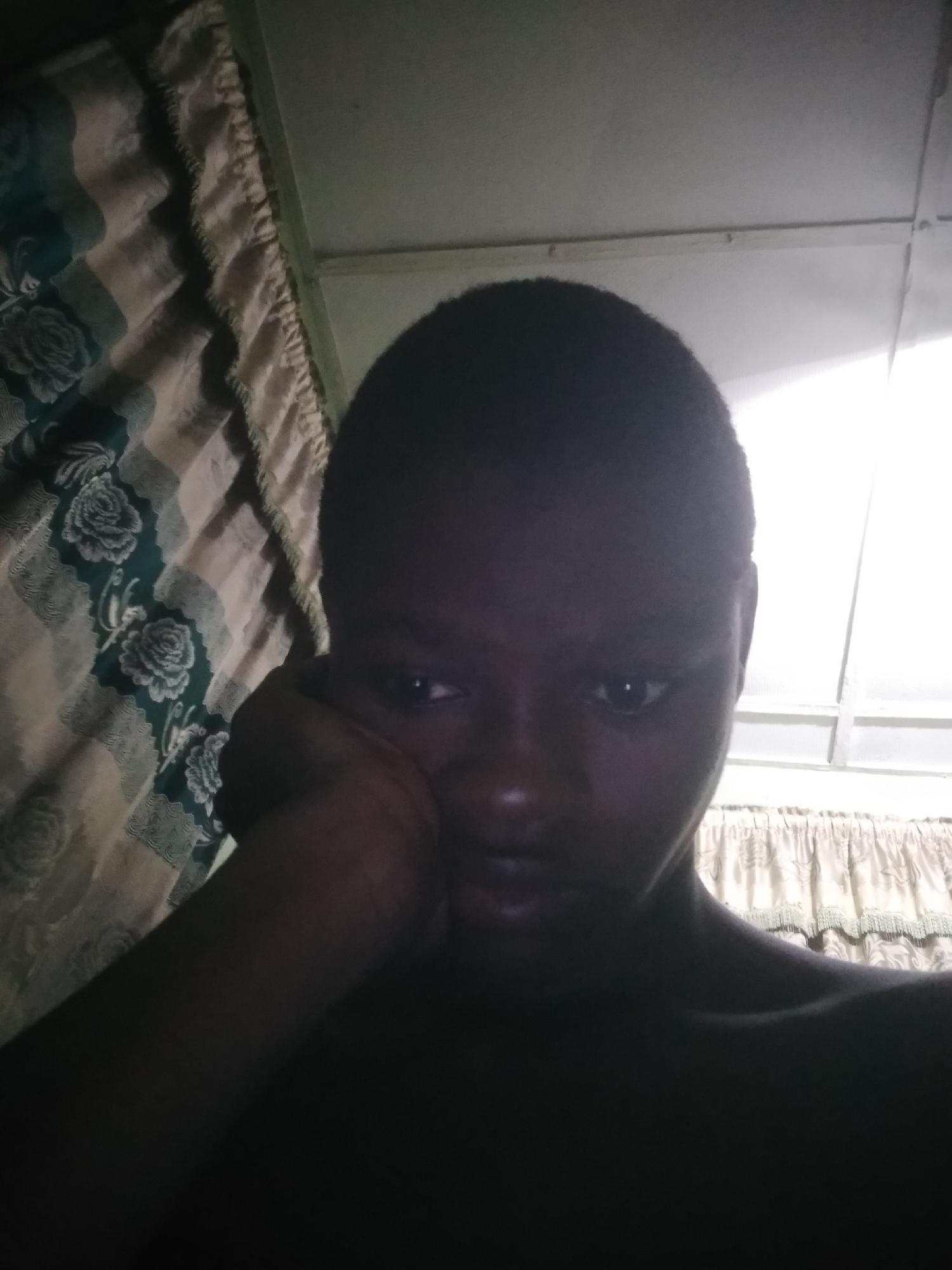 A person with dark skin and a shaved head is resting their head on their hand. The person appears to be indoors with curtains visible behind them. The lighting is poor, casting shadows on the person's face and upper body. The location is Unknown, Unknown.