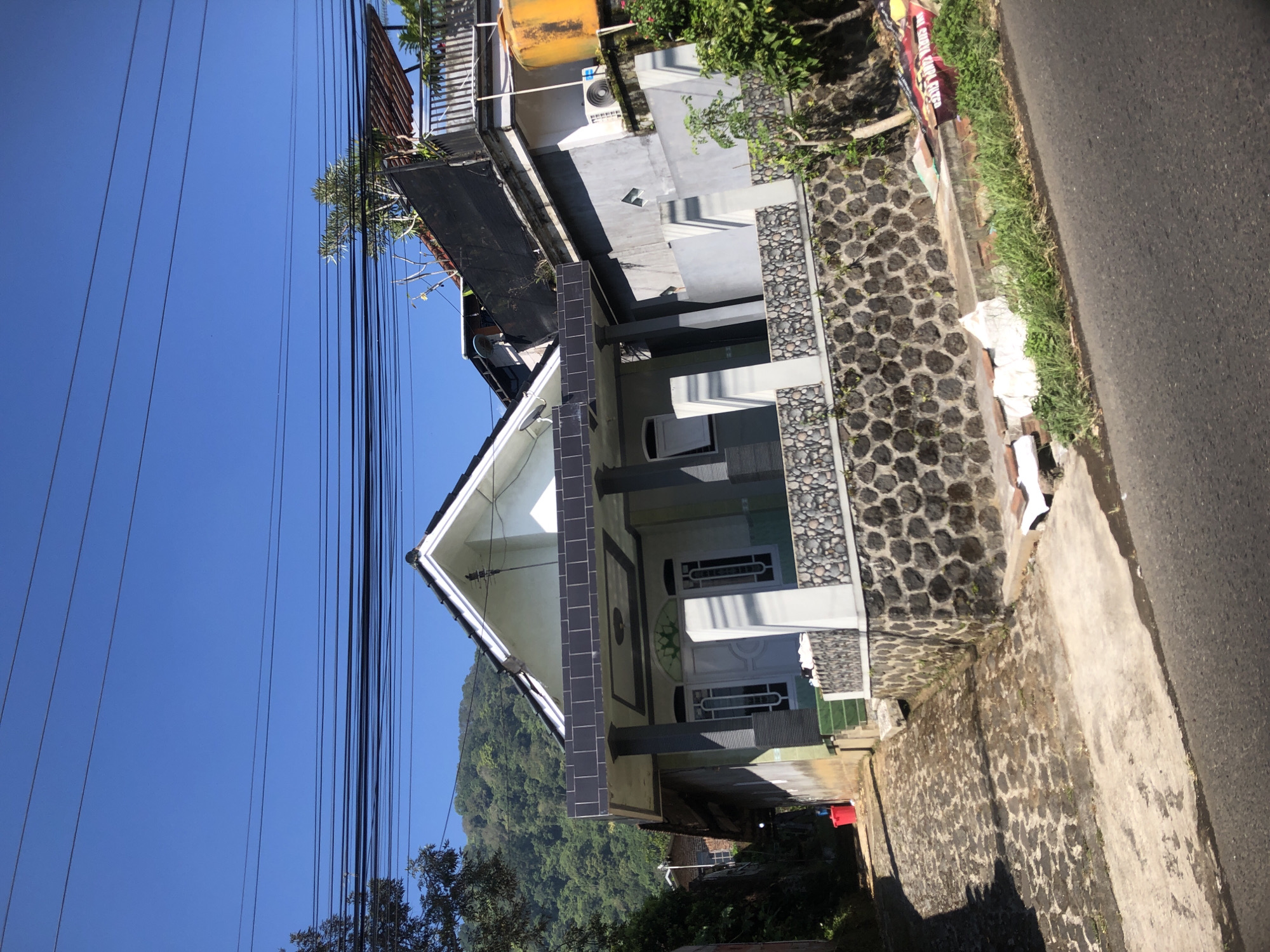 The image shows a street view in Trawas, Indonesia, under a clear blue sky. A two-story house with a modern facade is prominent on the right side of the street. The house features a white and grey exterior with dark tiled accents on the roof overhang. A stone wall with round decorative elements lines the front of the property. Electrical wires are strung across the top of the frame. The street itself is paved with cobblestones in the foreground, transitioning to asphalt further down. To the left of the house, a red bucket is visible. The background reveals a lush green hillside. A large yellow water tank is positioned on a neighboring structure to the right of the main house. Debris, including what appears to be cardboard and plastic, is scattered along the roadside in the lower right corner.
