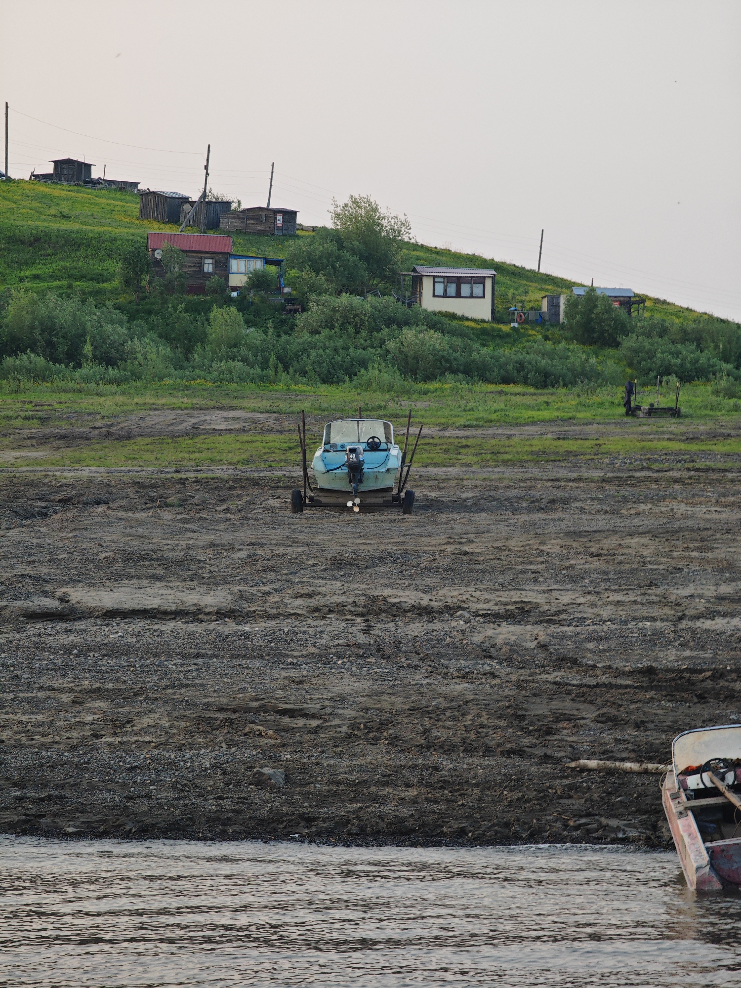 A wide shot captures a rural scene in Potapovo, Russia, on a clear day, likely in the late afternoon or early evening given the soft, diffused light. The foreground is dominated by a dry, gravelly riverbed, with the calm, rippling water of a river visible at the bottom of the frame. A reddish boat, partially submerged, rests at the edge of the river on the right side of the image.
Further back, a small, light blue motorboat is beached on the gravelly shore, resting on a trailer. It is positioned centrally in the frame, facing the viewer. A darker, distant figure stands on the grassy embankment behind the boat, engaged in an unseen activity.
The middle ground features a lush green, sloping hillside dotted with small yellow flowers and scattered trees. Along the crest of the hill, a cluster of simple wooden houses and structures are visible, suggesting a small settlement. Power lines stretch between poles, connecting the dwellings. The architecture of the houses, with their pitched roofs and wooden construction, indicates a rural or remote setting. The overall atmosphere is one of quiet, everyday life in a northern landscape. No visible text is present in the image.