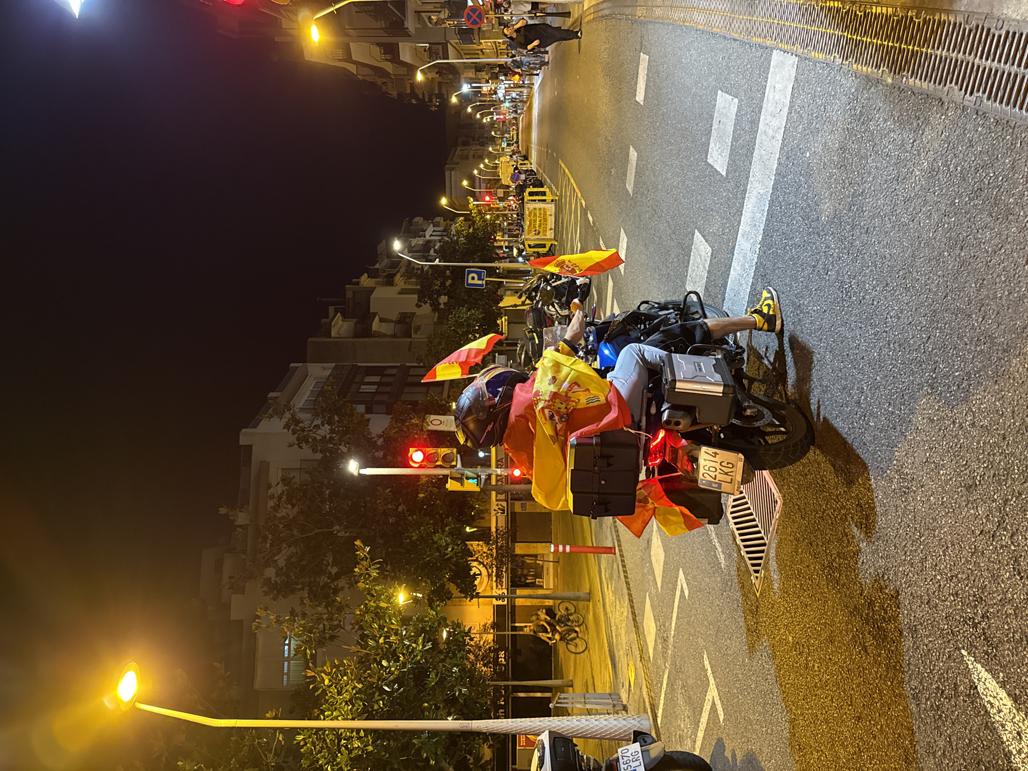 A nighttime street scene in Blanes, Spain, features a person on a motorcycle adorned with Spanish flags. The motorcycle, a BMW R1250GS with license plate 2614 LKG, is stopped at a pedestrian crossing. The rider, wearing a helmet and yellow and black sneakers, is draped in Spanish flags. In the background, two people are riding bicycles on the sidewalk. The street is lined with buildings and illuminated by streetlights and traffic signals. The atmosphere is festive, possibly due to a local event or celebration.